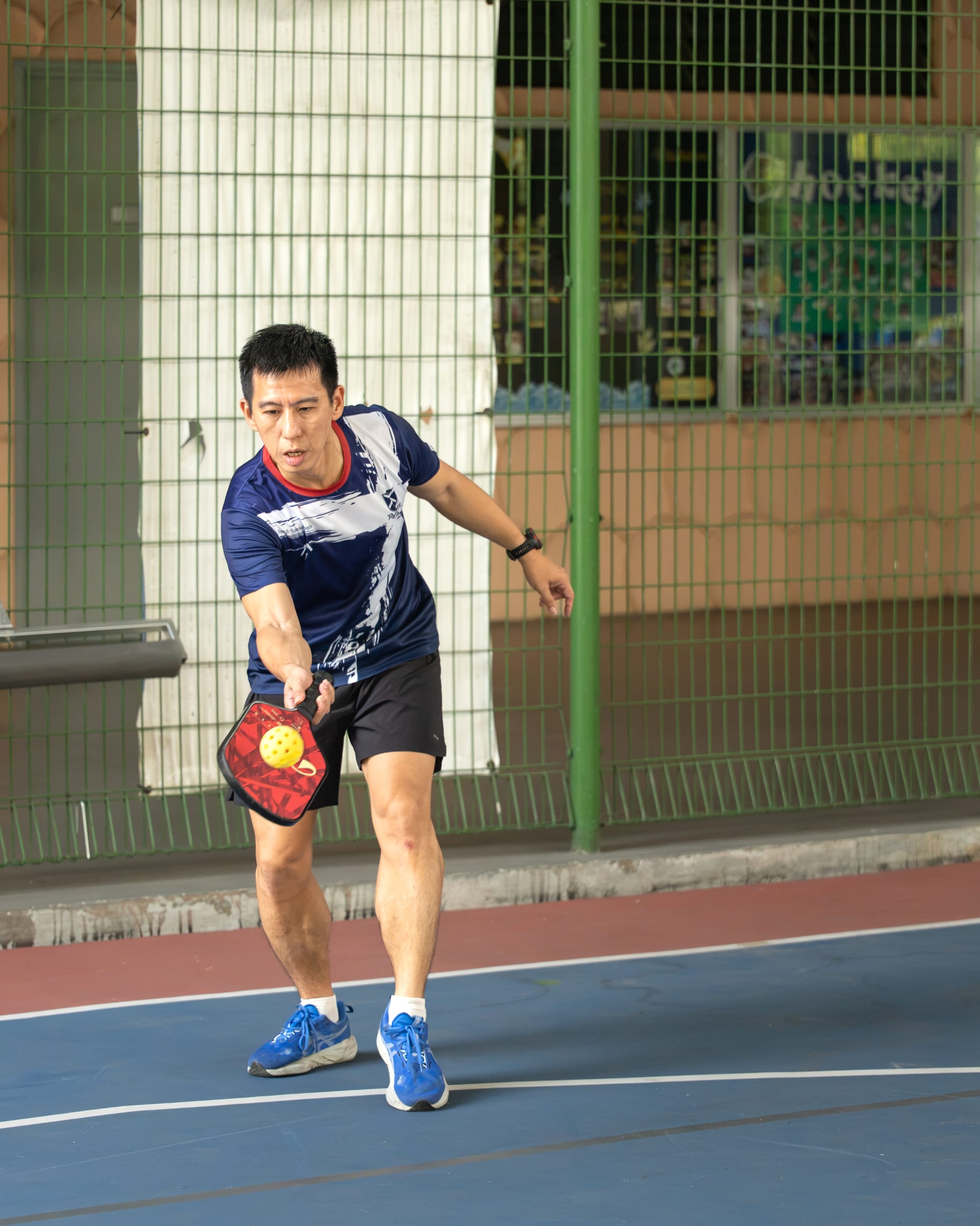 A focused man in a navy blue graphic t-shirt, dark shorts, and blue sneakers mid-action on a court. He leans low to strike a yellow perforated pickleball with a red-patterned paddle, set against a background of a green fence and building windows.