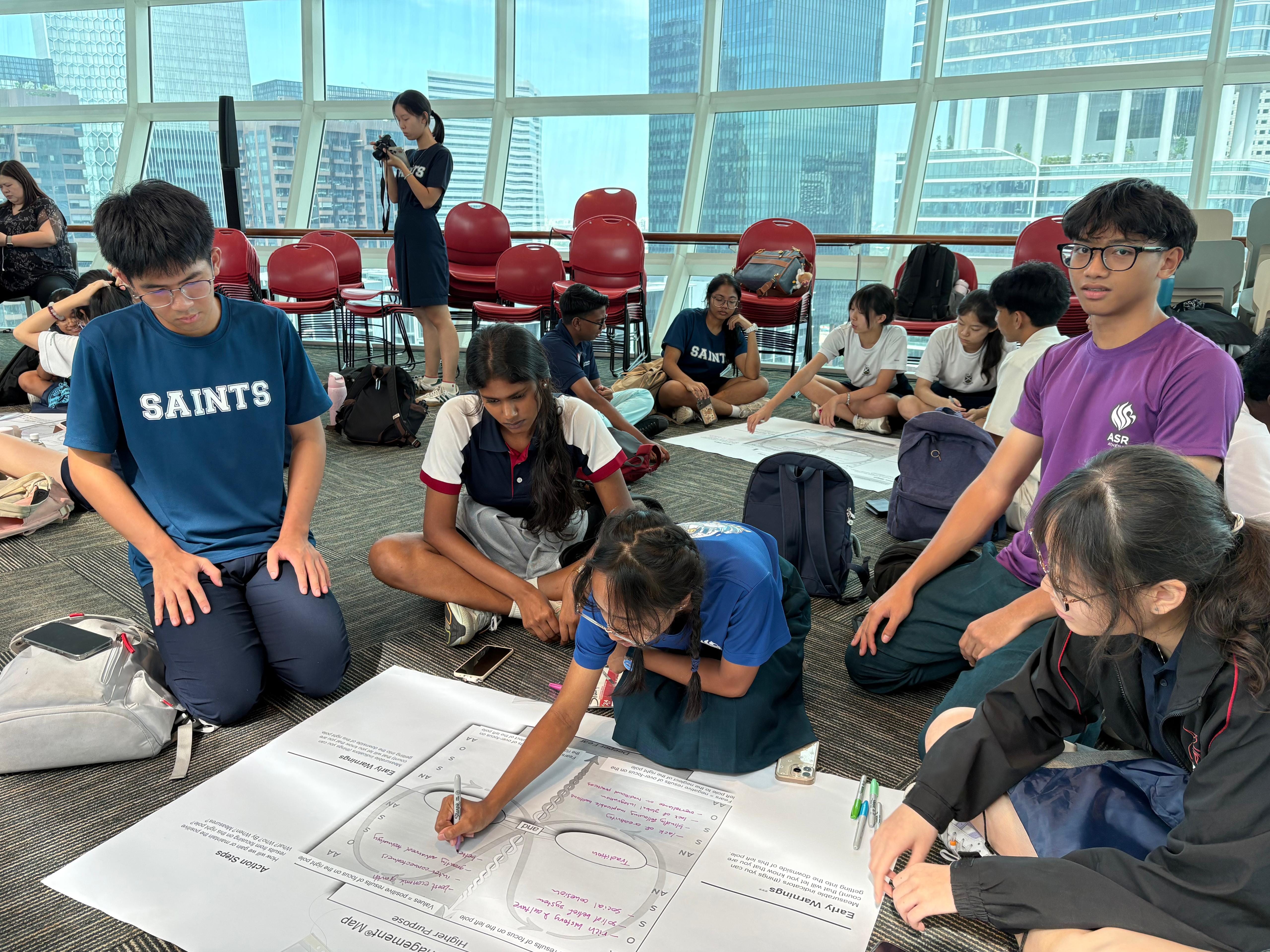 Students from various schools sit on the floor in a spacious, well-lit room, working in small groups on large printed activity sheets, with a city skyline visible in the background.