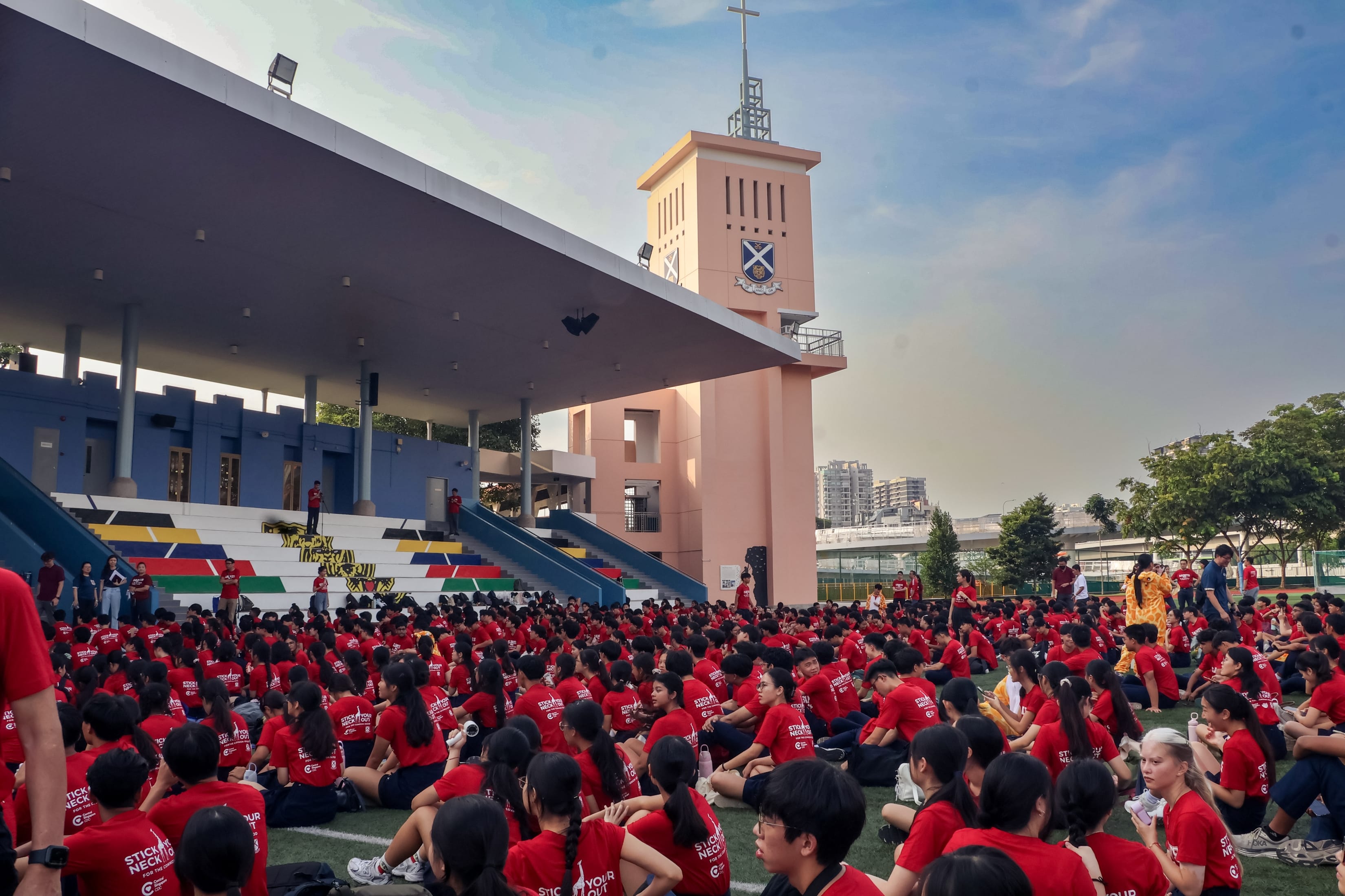 A large crowd of students in red t-shirts sitting outdoors on a field in front of a grandstand and a tall peach-colored tower topped with a cross.
