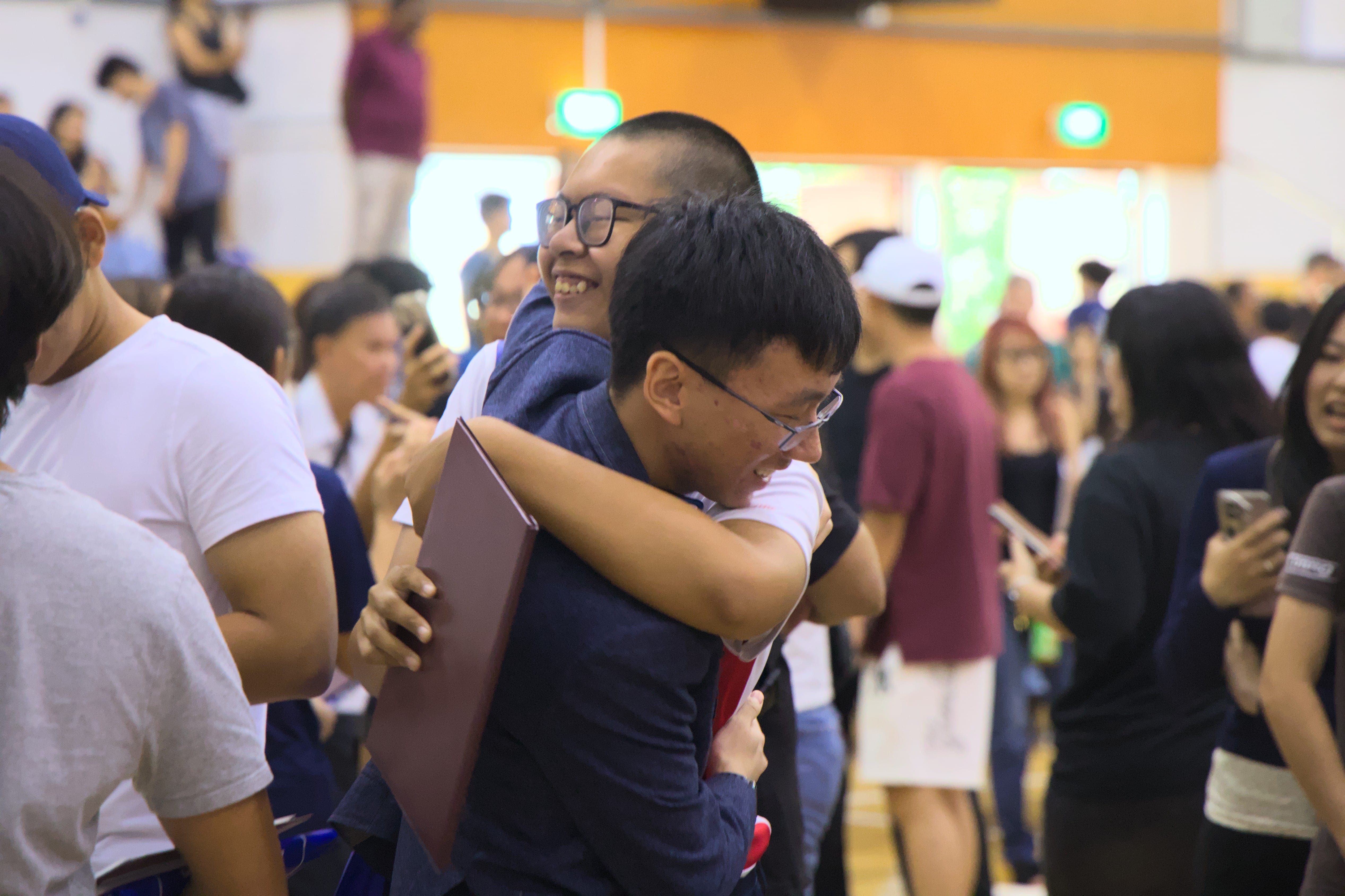 Two young men wearing glasses hug and smile in a crowded indoor hall with many people in the background.