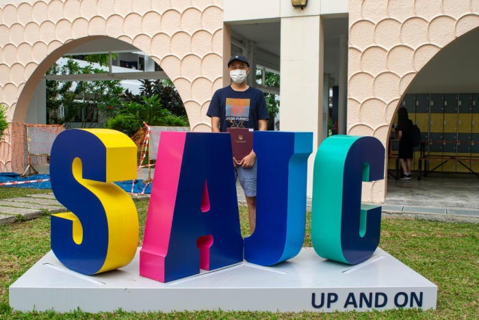 A masked man stands behind a colorful SAJC sign reading "UP AND ON" outside a building.