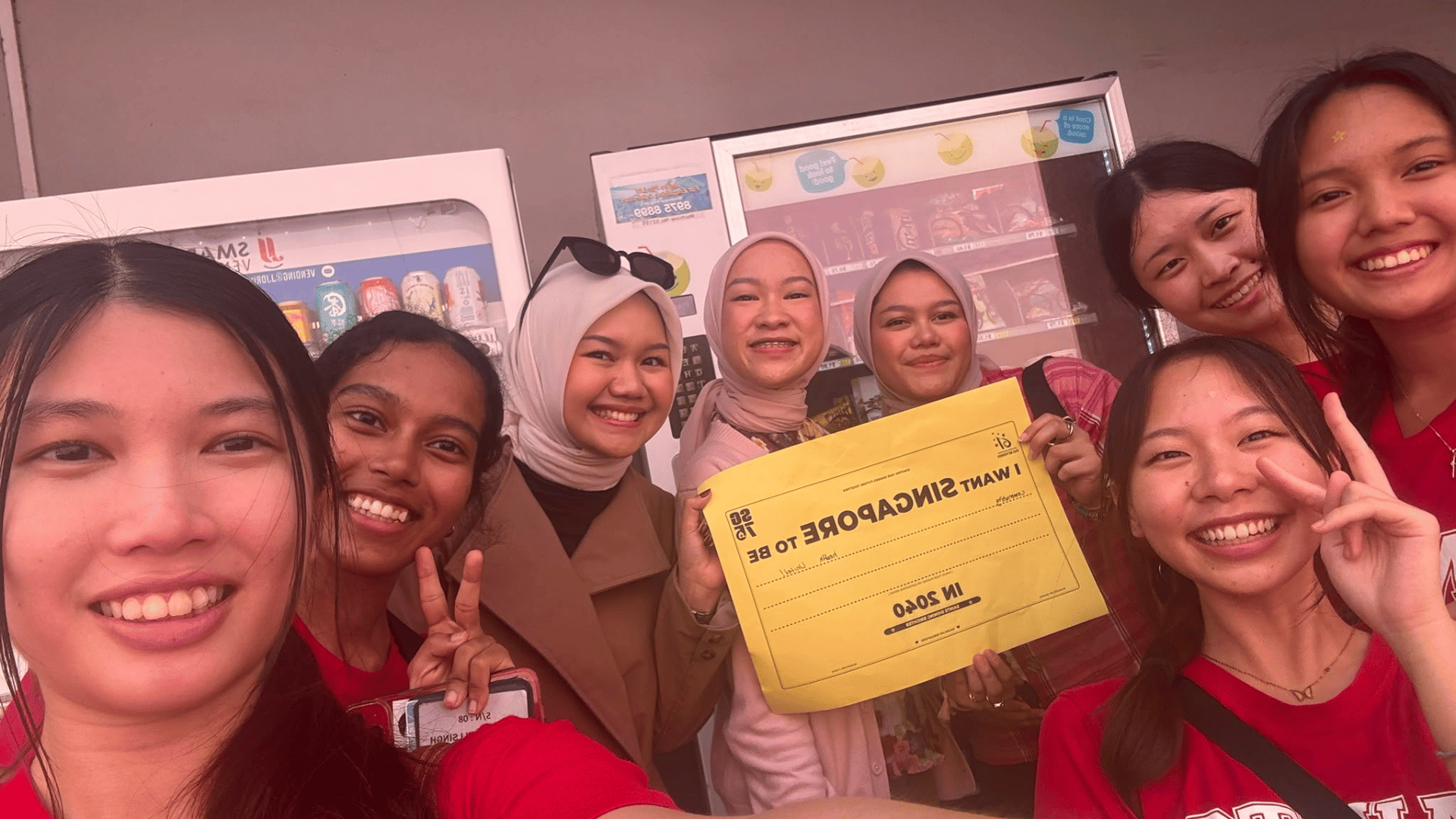 A selfie of a diverse group of smiling young women gathered closely outdoors in front of white beverage and snack vending machines. Near the center, a woman holds up a yellow paper sign with upside-down printed text reading "I WANT SINGAPORE TO BE... IN 2050." Several of the women are dressed in red t-shirts, two wear headscarves, and some flash peace signs toward the camera.