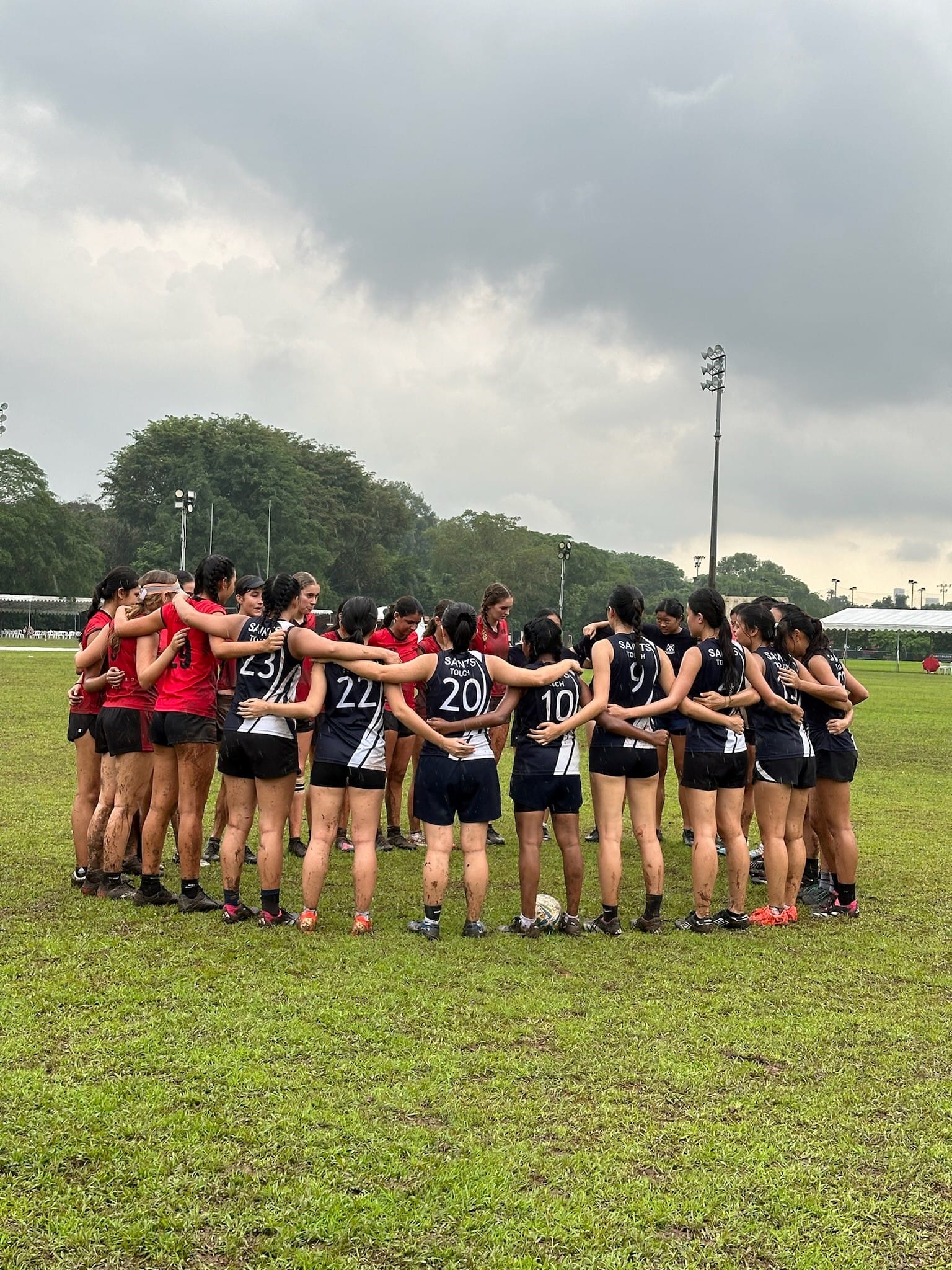 Players from two touch rugby teams - one in black and white 'Saints Touch' jerseys and one in red - huddle together with arms around each other on a muddy grass field after a match, under an overcast sky.