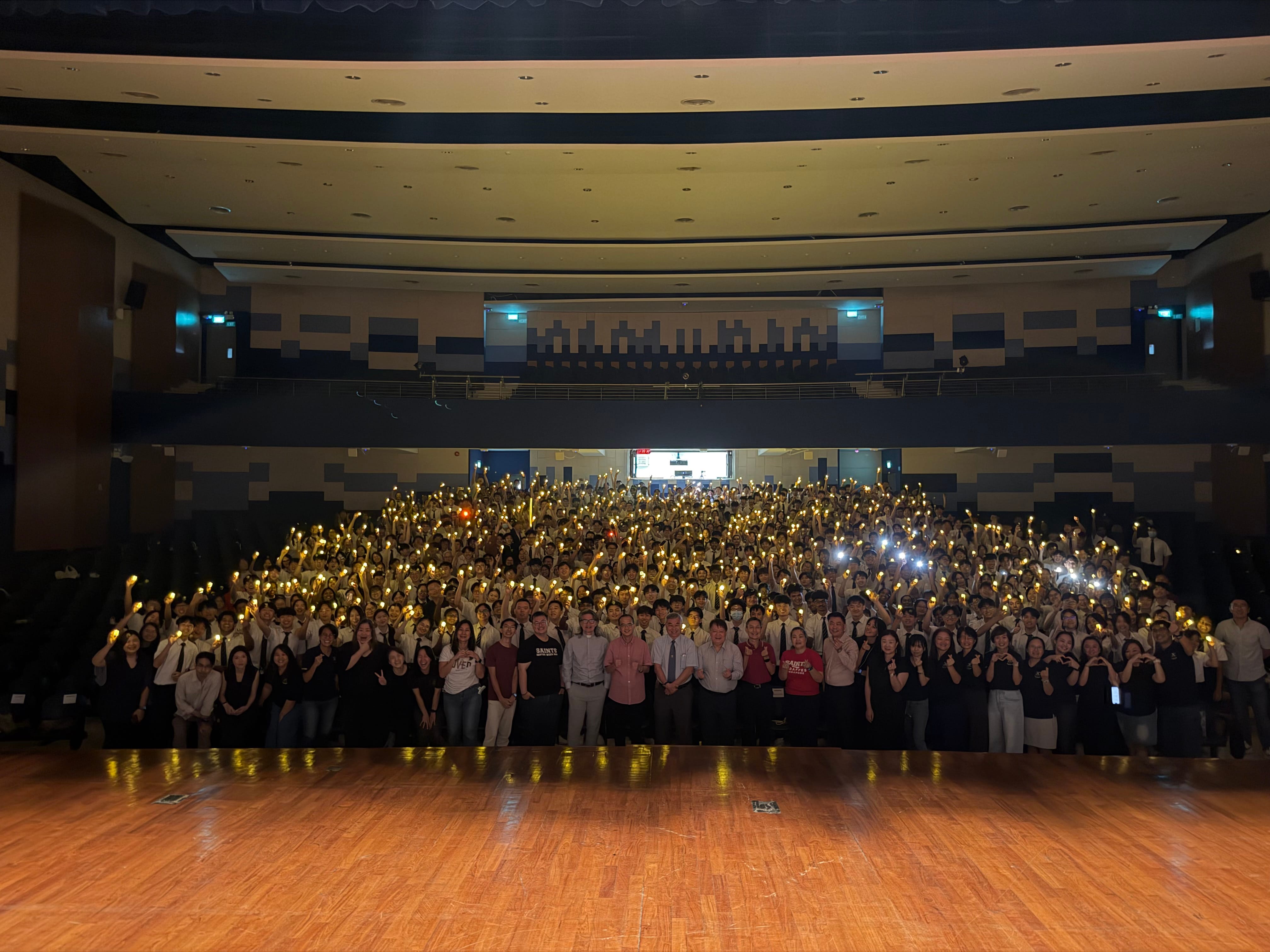 A large group of students and staff gathered in an auditorium, viewed from the wooden stage, holding up small glowing lights or LED candles.