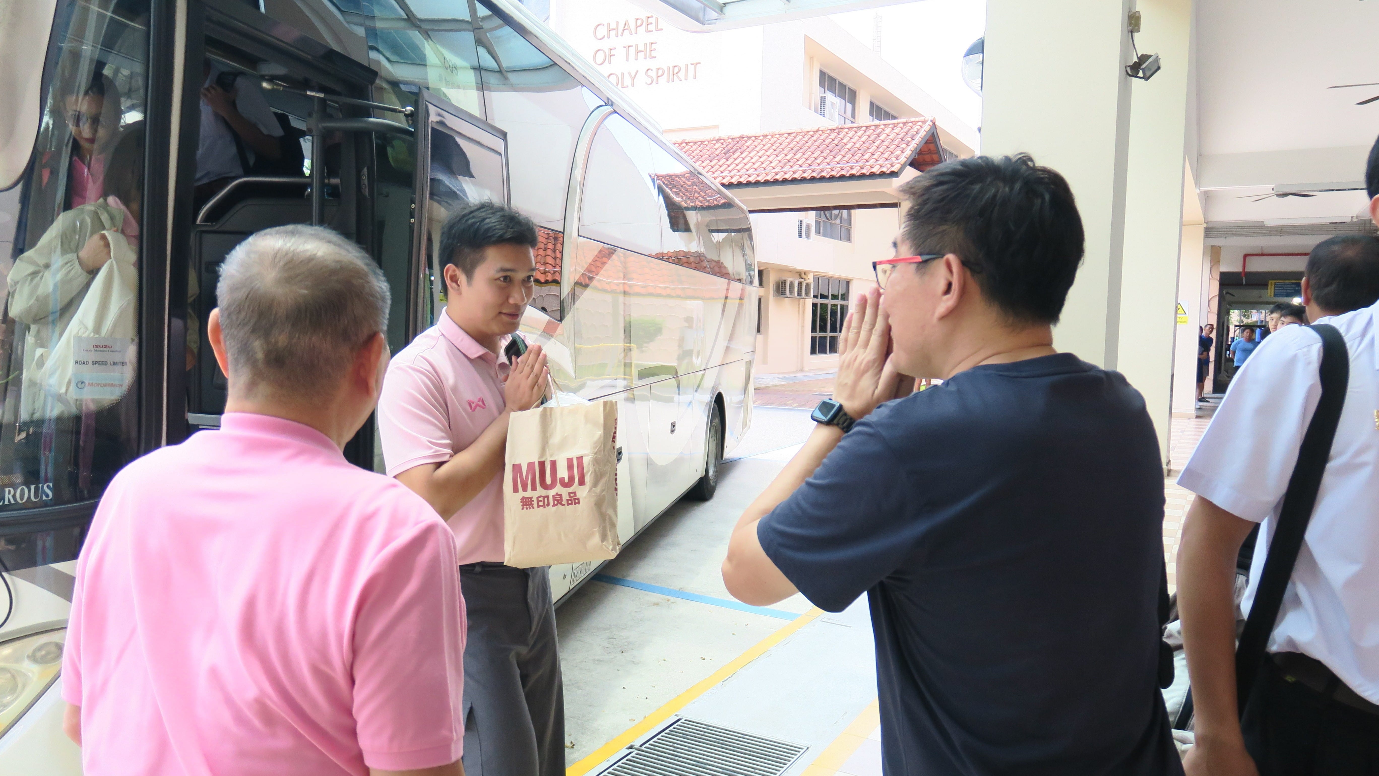 A teacher in a pink polo shirt greets visitors beside a coach bus outside a school building, with a "Chapel of the Holy Spirit" sign visible in the background.