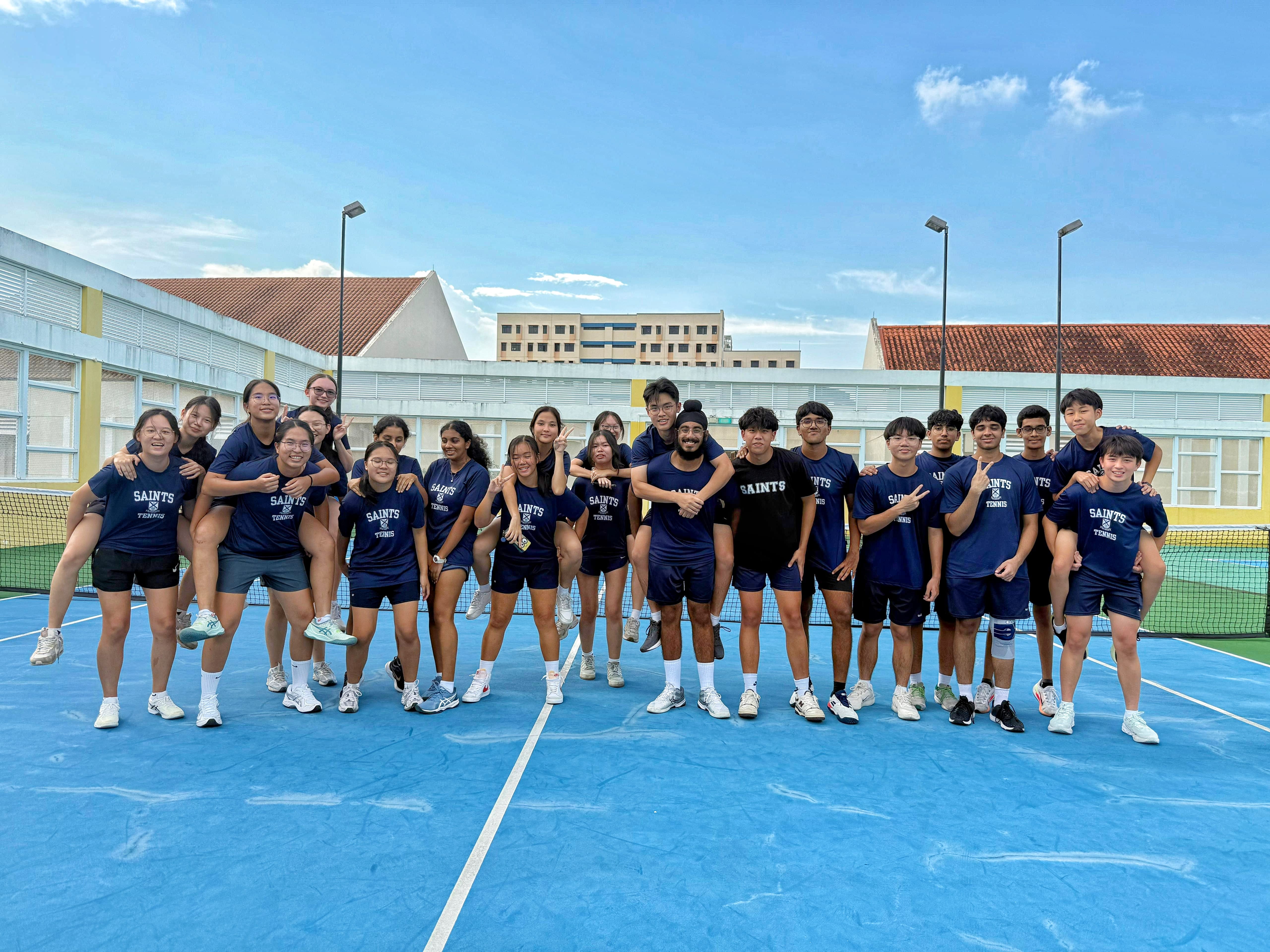 A large group of Saints Tennis players in matching navy blue 'Saints Tennis' t-shirts pose cheerfully together on a blue outdoor tennis court, with school buildings visible in the background under a bright blue sky.