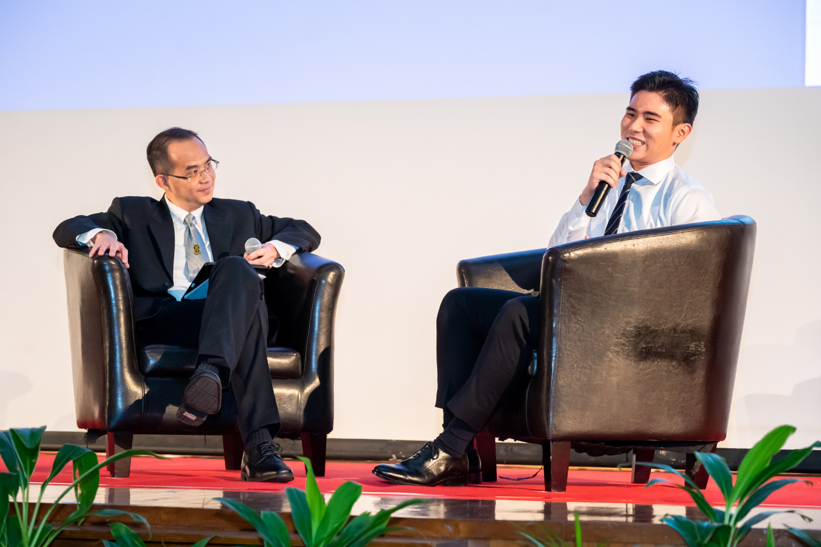 Mr Tham with valedictorian seated on the stage with a red carpet, engaged in what appears to be a interview-style conversation.