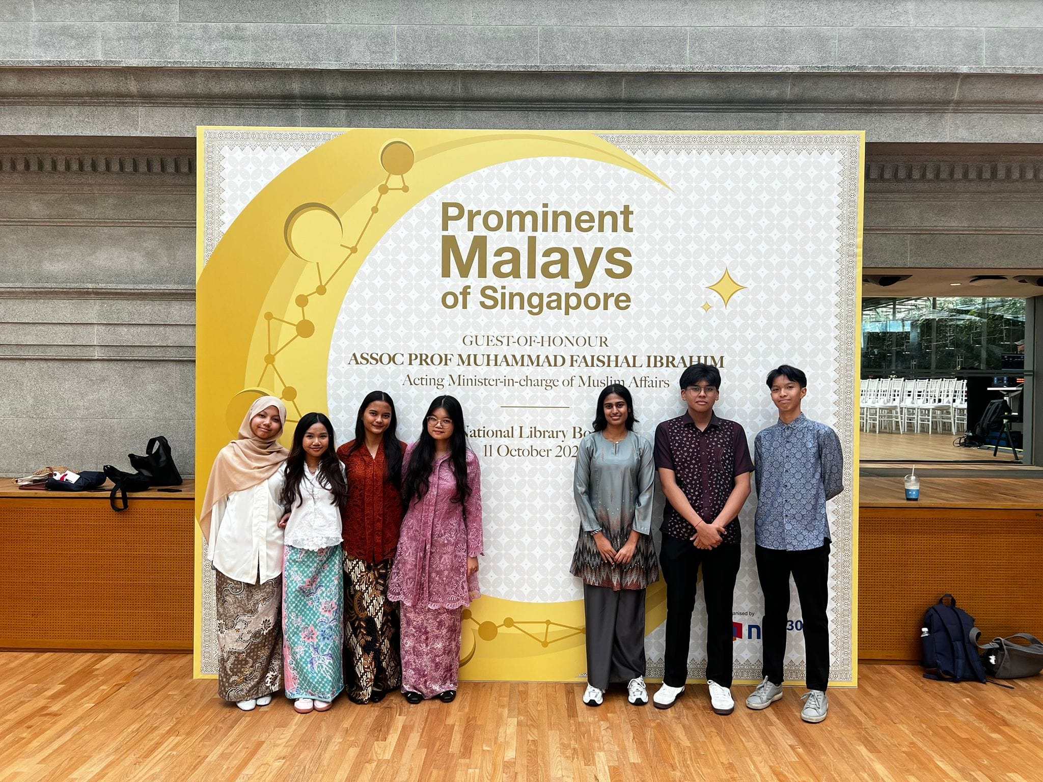 SAJC student volunteers dressed in traditional attire posing in front of the 'Prominent Malays of Singapore' event backdrop at the National Library Board, organised on 11 October 2025 with Guest-of-Honour Assoc Prof Muhammad Faishal Ibrahim, Acting Minister-in-charge of Muslim Affairs
