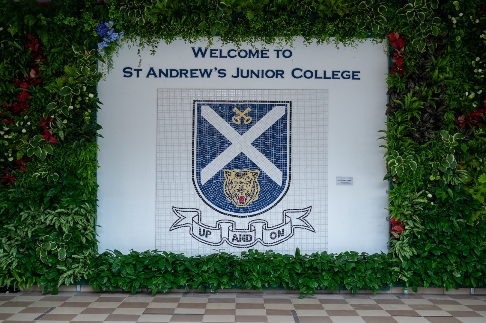 White wall with Welcome to St Andrew's Junior College sign and mosaic crest, surrounded by a lush green plant wall.