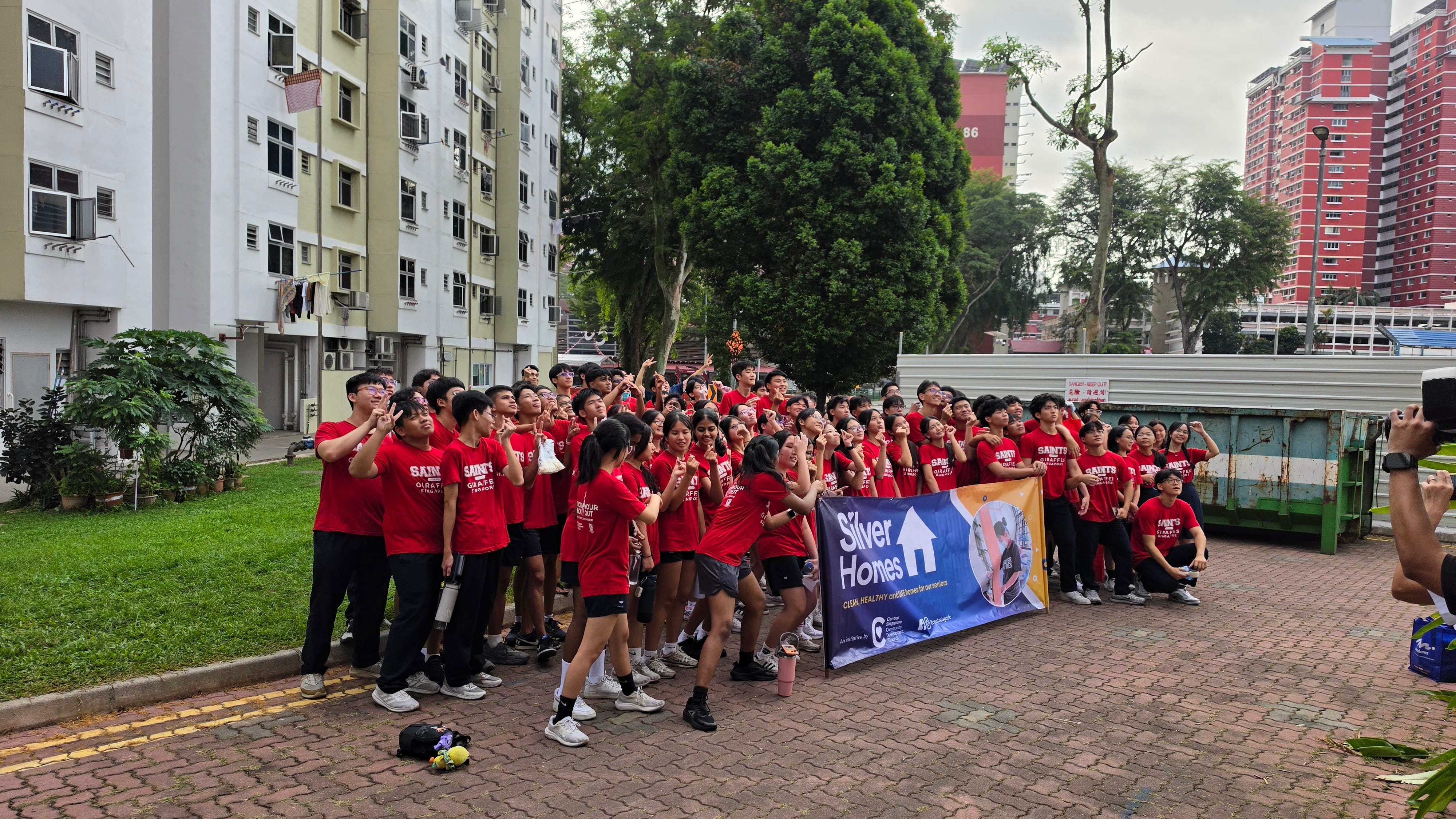 A group of student volunteers dressed in matching red "SAINTS" t-shirts gather outdoors on a paved courtyard next to a grassy area in a residential neighborhood. They smile and gesture cheerfully while holding a large blue banner that reads "Silver Homes" with the tagline "Clean, Healthy environments for seniors." High-rise residential housing blocks and green trees surround the area in the background.