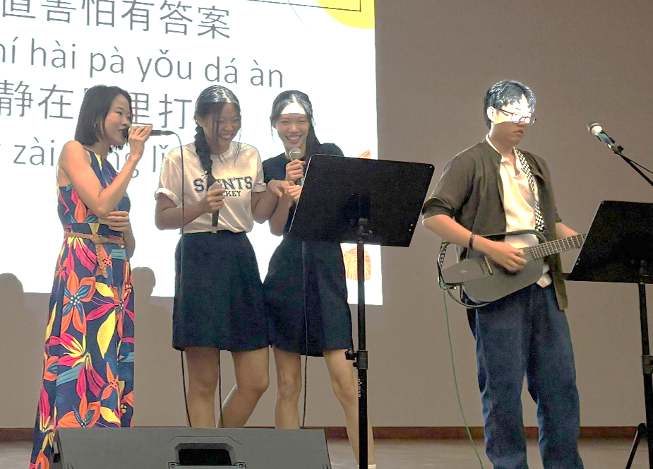 A performance on stage where a teacher in a floral dress and two students sing into microphones, accompanied by a student playing a ukulele. Chinese lyrics with pinyin are projected on the screen behind them.