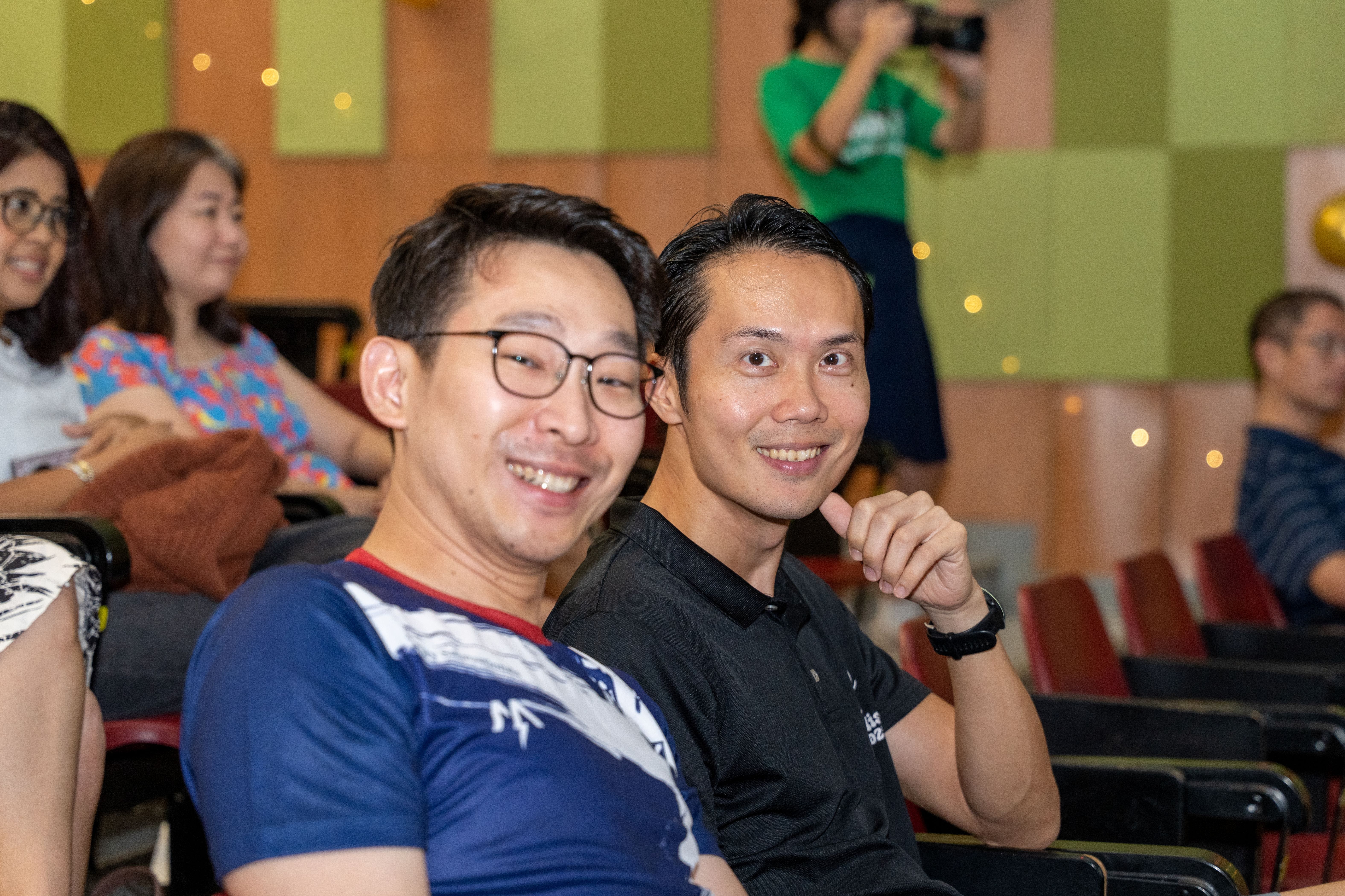 A medium close-up of two men seated side-by-side in auditorium chairs. The man on the left wears glasses and a navy blue patterned t-shirt while smiling broadly, while the man on the right wears a black polo shirt and smiles directly at the camera. Other seated audience members and a person taking a photo in a green t-shirt are visible in the background.