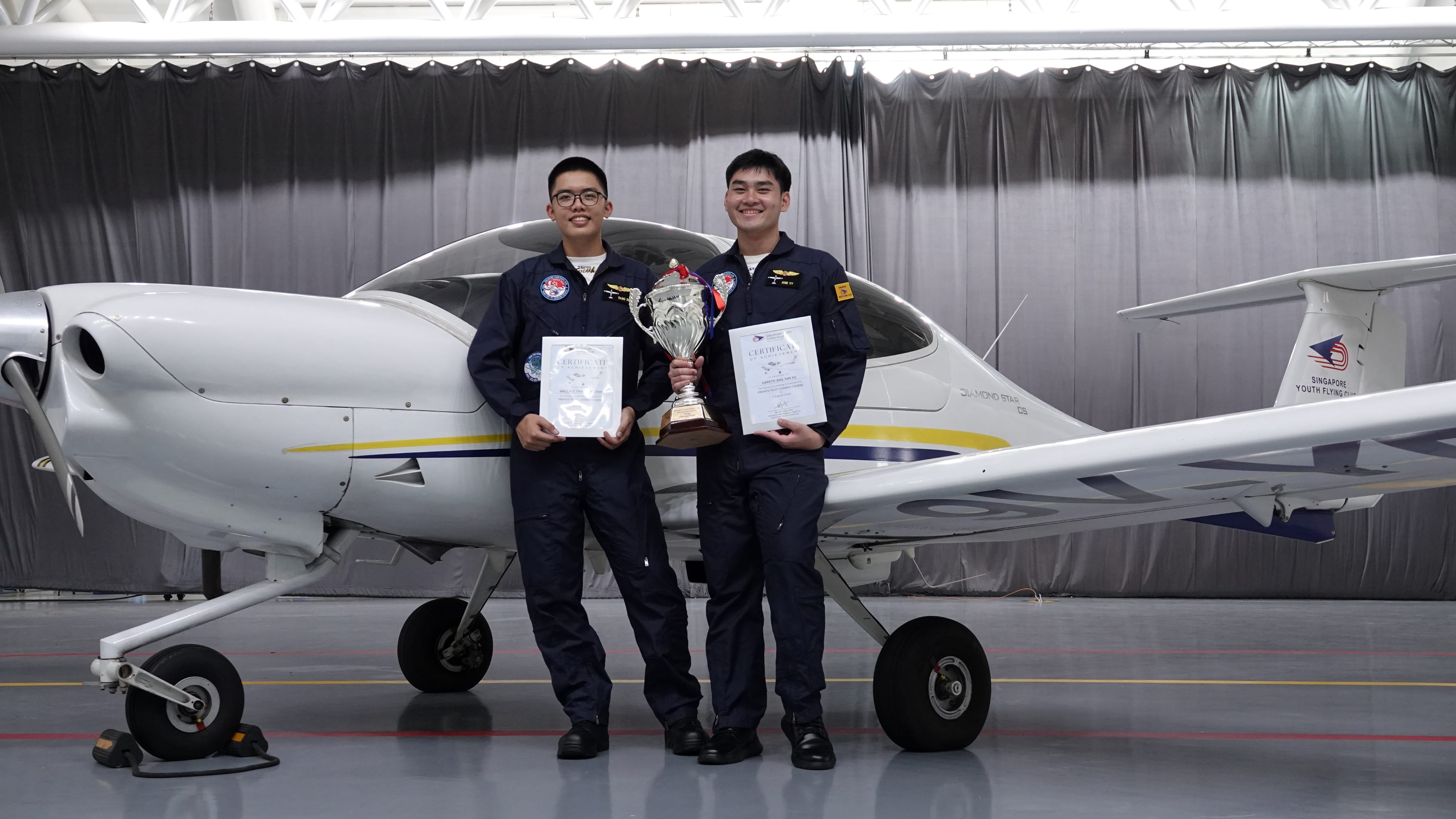 Two young men in navy flight suits hold certificates and a trophy while posing in front of a Diamond Star aircraft inside a hangar belonging to the Singapore Youth Flying Club.