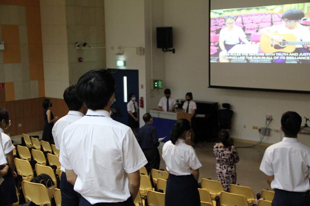 Students in school uniforms stand facing a large projection screen in an auditorium during what appears to be a worship or assembly session, with lyrics displayed on the screen.