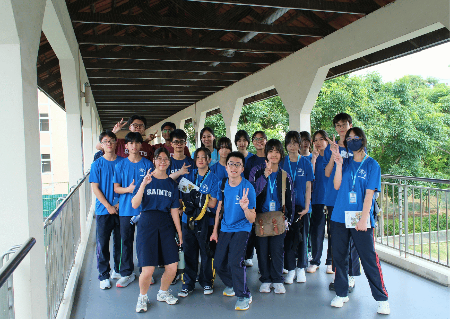 A cheerful group of students in blue T-shirts from Tsung Tsin Christian Academy and St Andrew's Junior College pose together along the Jacob Ballas Bridge, many flashing peace signs, with lush greenery visible in the background.