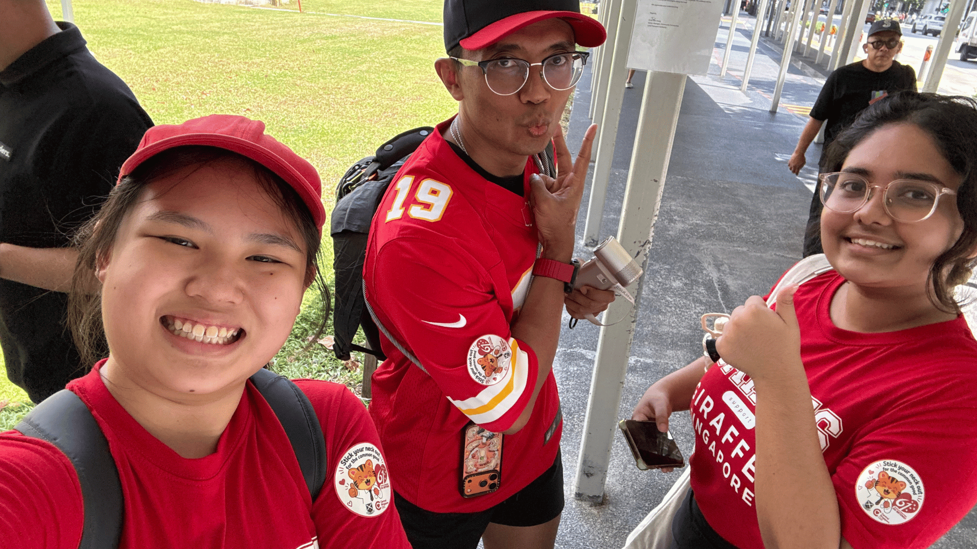 A close-up selfie of three individuals outdoors on a sheltered walkway next to a grass lawn. On the left, a smiling young woman wears a red cap and red t-shirt. In the center, a man wearing glasses, a red cap, and a red sports jersey with the number 19 makes a playful face and peace gesture. On the right, a young woman in glasses and a red t-shirt gives a thumbs-up. Each person wears a round event sticker on their sleeve.