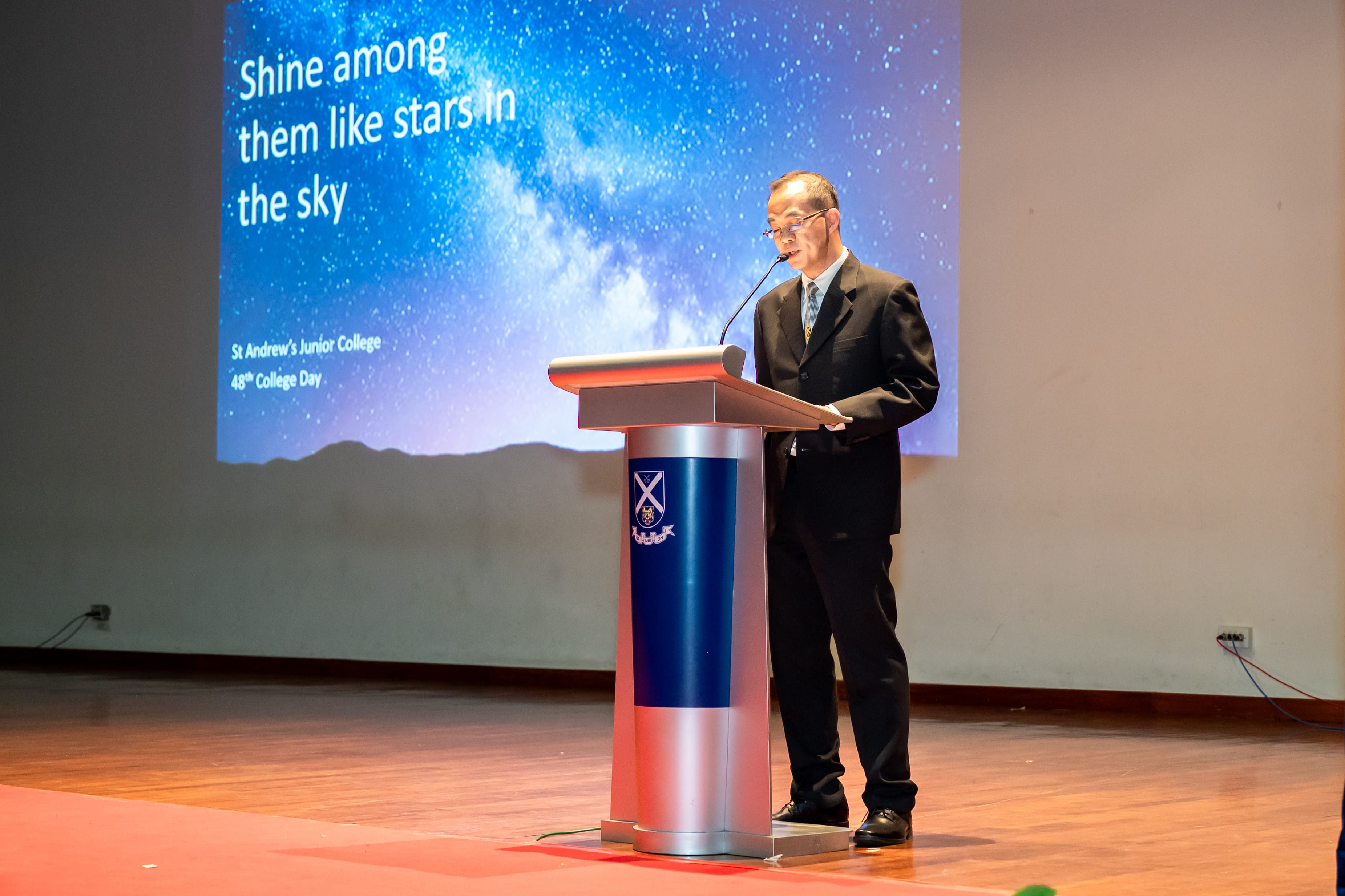 Man in a suit speaks at a podium onstage, with a starry sky slide reading Shine among them like stars in the sky.