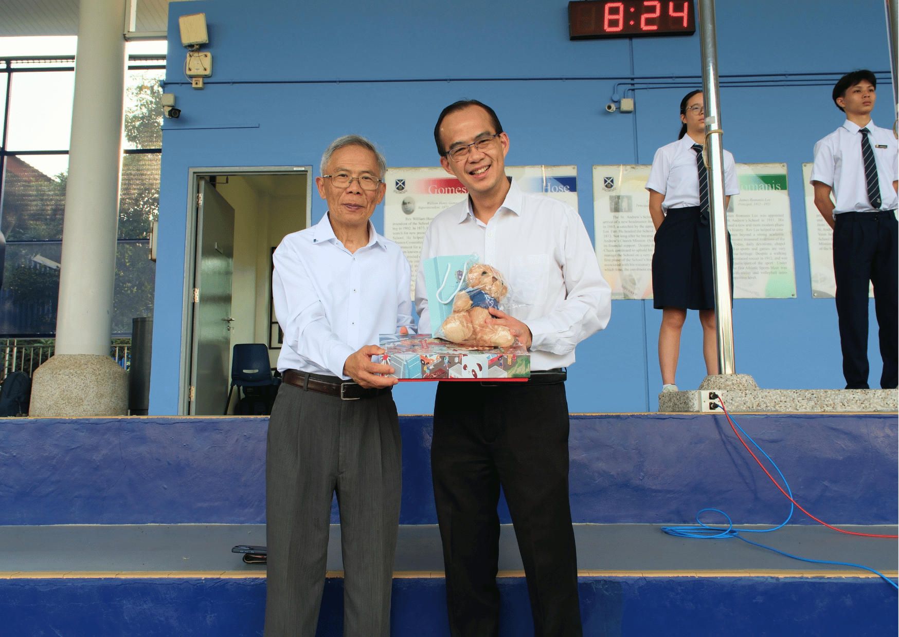 Mr Cheung, Principal of Tsung Tsin Christian Academy, presenting a gift set including a teddy bear to Mr Tham, Principal of St Andrew's Junior College, with students standing in the background.