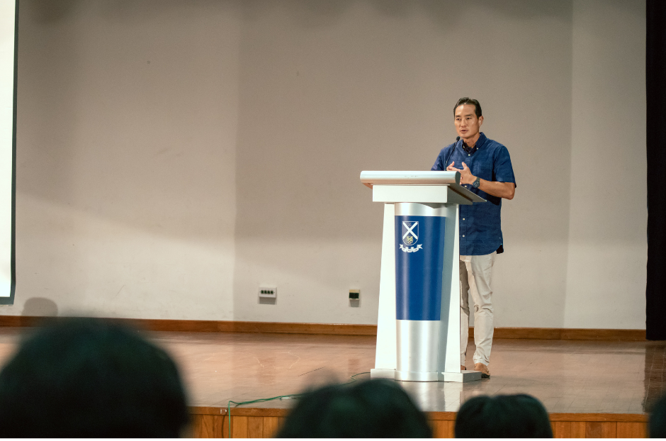 SMS Mr Tan Kiat How addresses an audience from a white podium bearing the St Andrew's Junior College crest, on a stage in an auditorium.