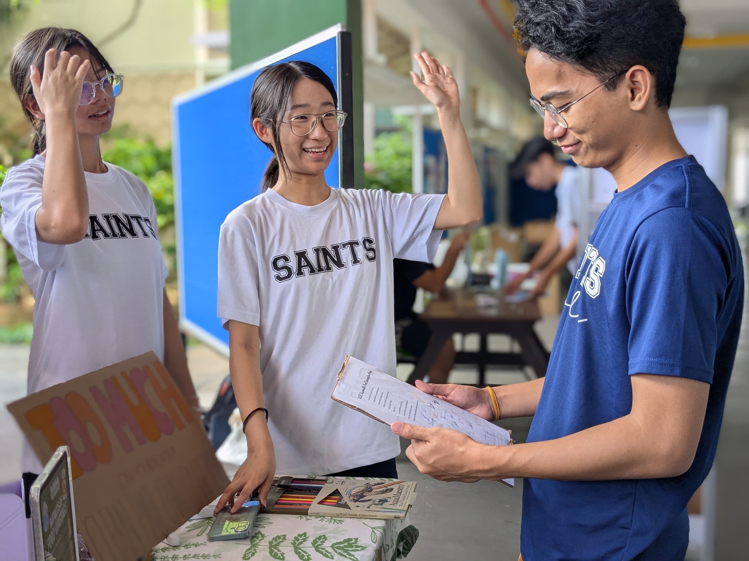 Two students in SAINTS shirts greet a student holding papers at a table with books and colored pencils.