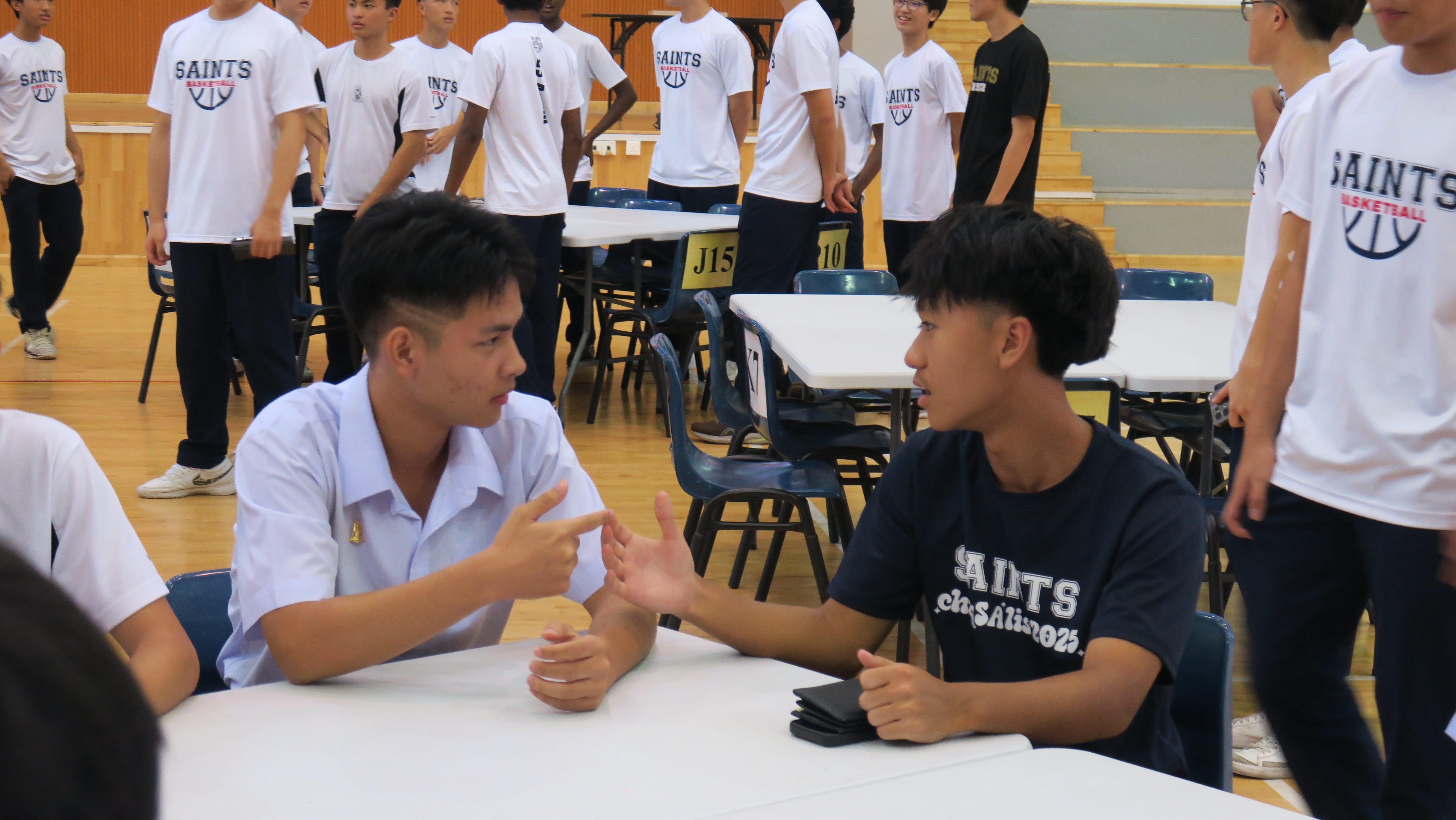 Two students — one in Thai school uniform and one in a Saints T-shirt — engage in an animated conversation at a table in a school hall, with other students in Saints Basketball jerseys standing in the background.