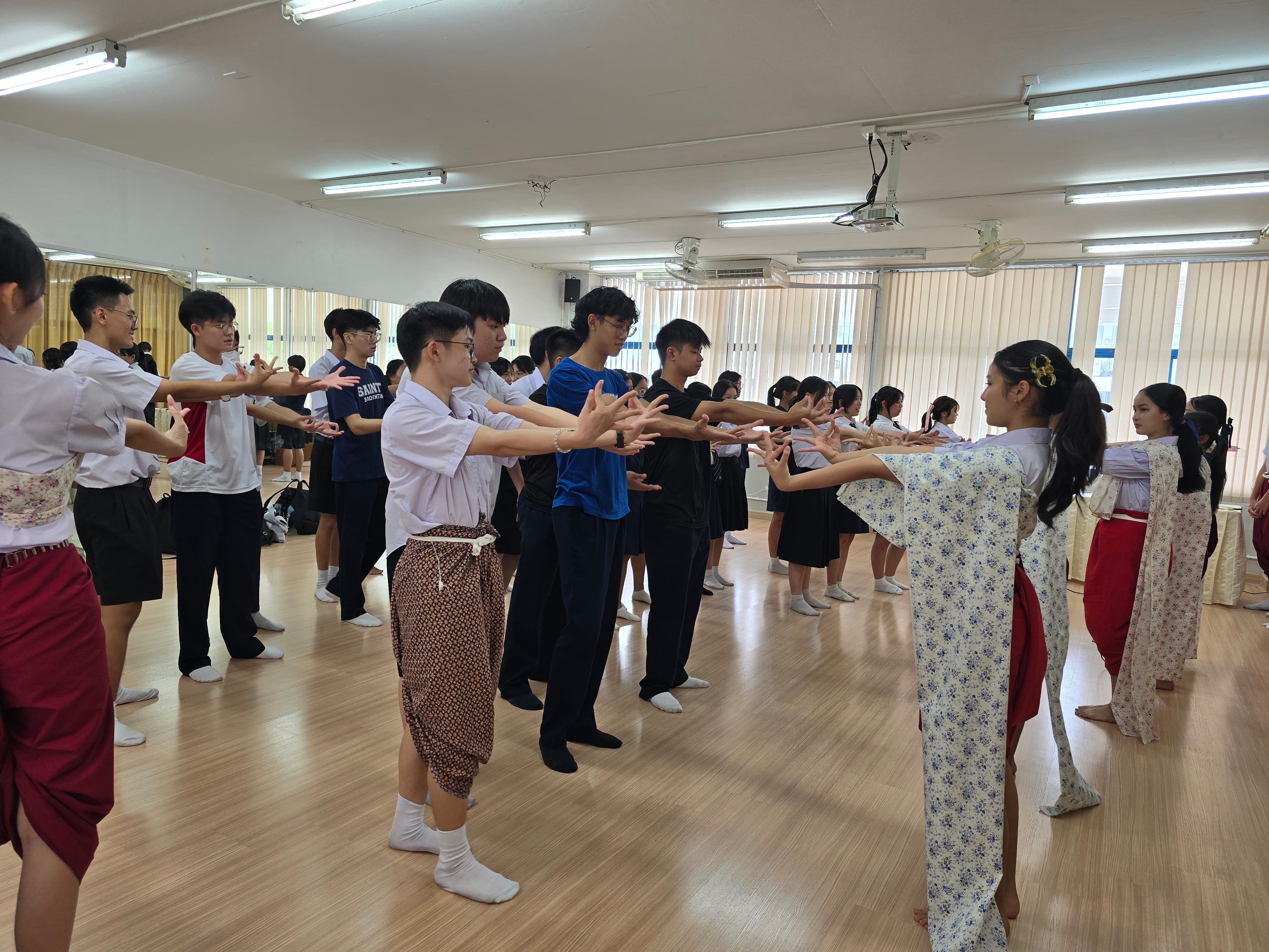 Students from St Andrew's Junior College and their Thai counterparts stand in two facing rows in a dance studio, practising traditional Thai dance hand gestures under the guidance of instructors in traditional Thai costume.