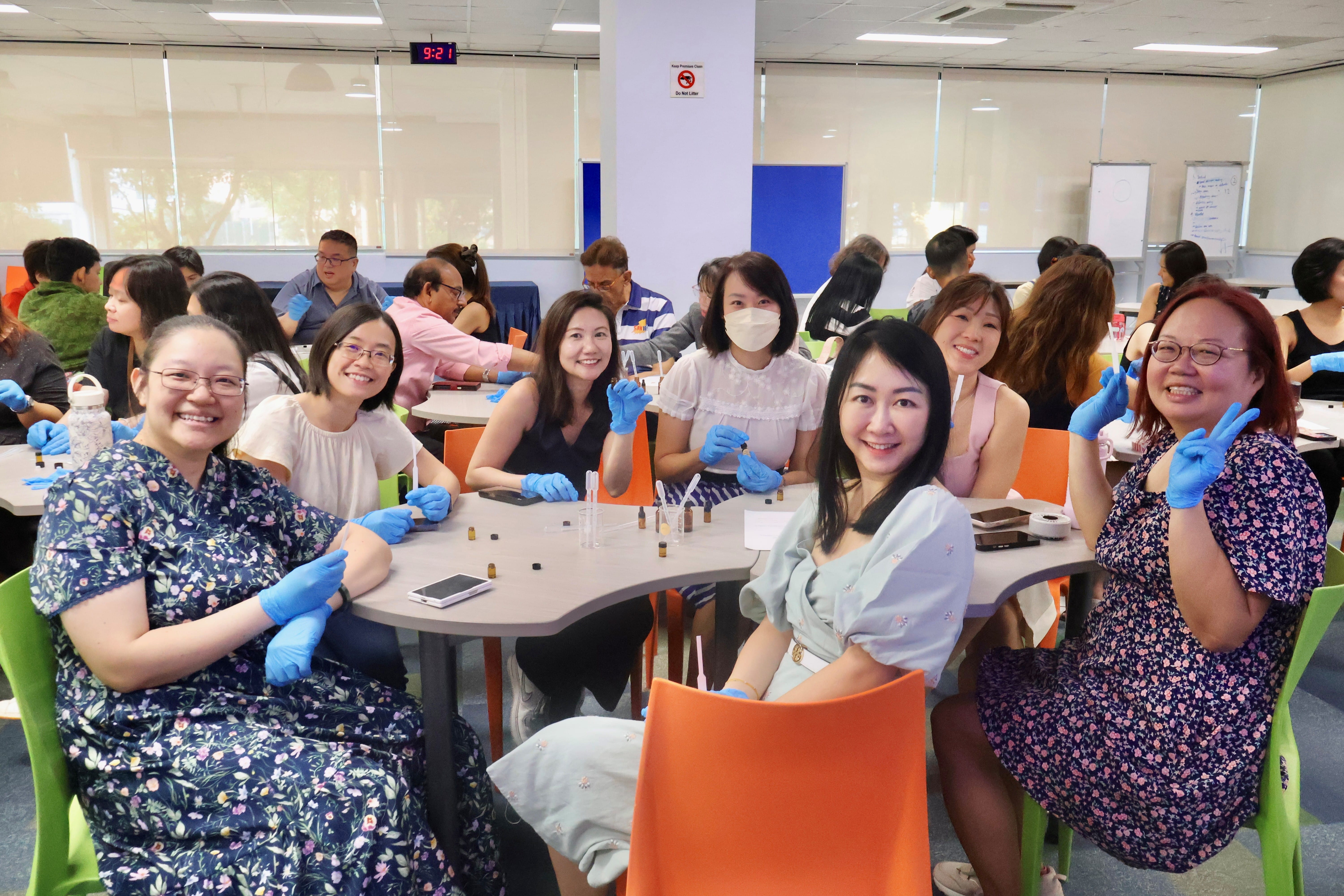 A group of women sit around a light-grey modular table in a brightly lit room, smiling together at the camera. Several attendees wear blue protective gloves and work with small glass dropper bottles on the table. The woman on the far right in a dark floral dress flashed a peace sign, while other participants work in groups in the background.