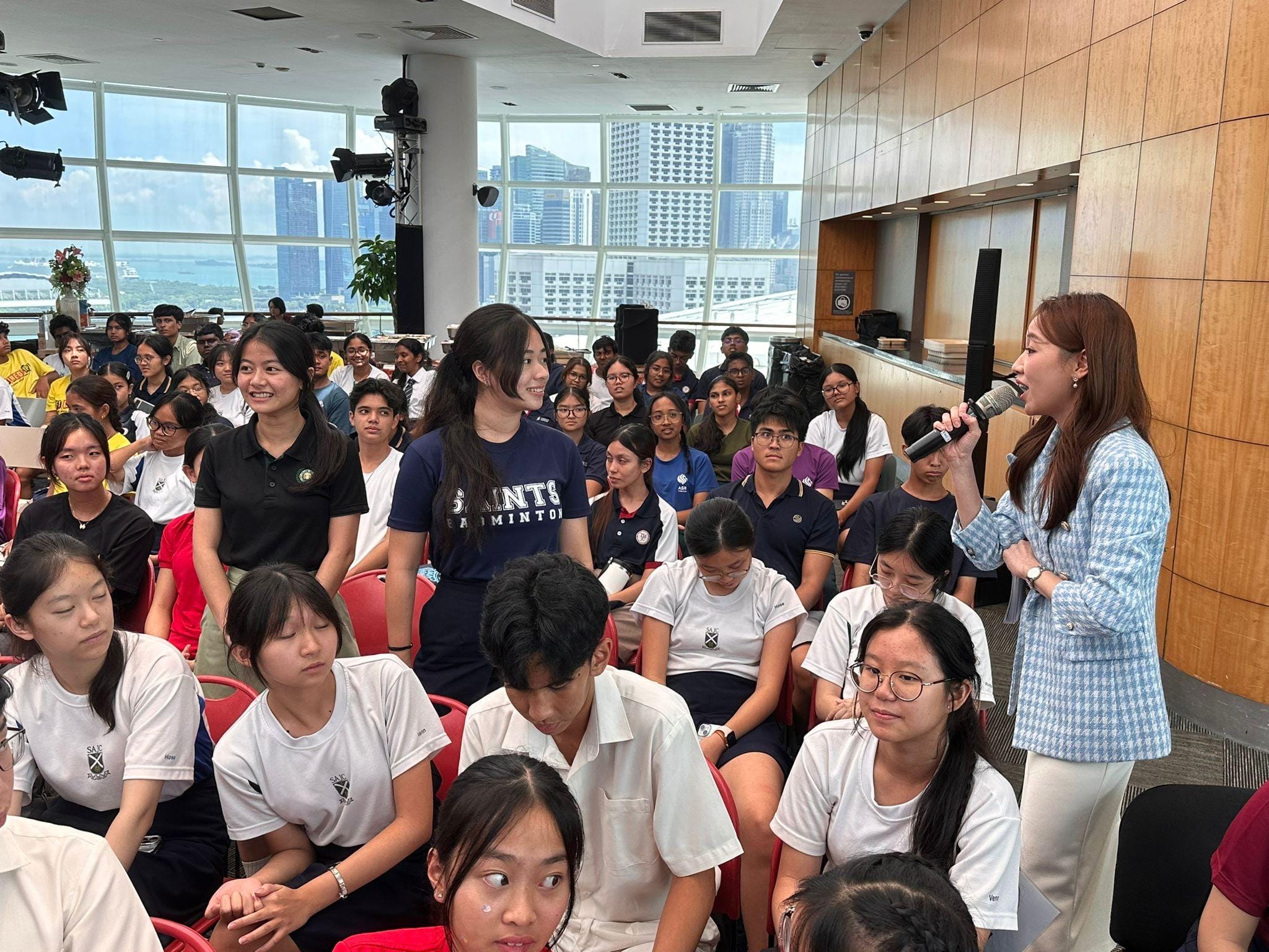 Our keynote speaker, Ms Tan Qin Lin from Mediacorp, speaks to a large group of students in school uniforms in a high-rise room with city views.