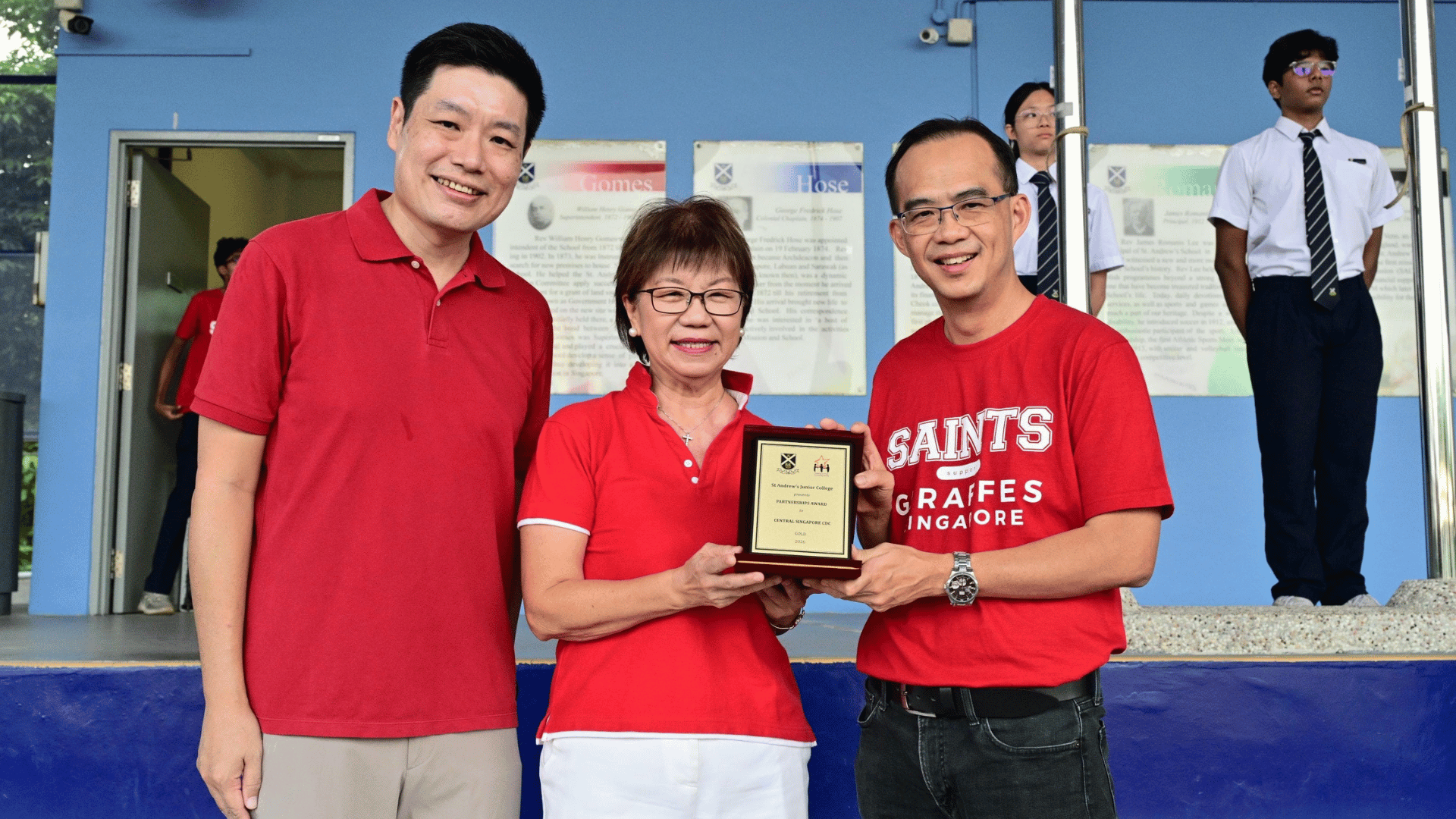 Mayor of Central Singapore CDC, Ms Denise Phua, Grassroots Advisor for Potong Pasir, Mr Alex Yeo and Principal of SAJC, Mr Tham Kine Thong, standing together in red tops on a stage in front of a blue wall with framed commemorative plaques. Mayor Phua and Mr Tham on the right wearing a red "SAINTS GIRAFFES SINGAPORE" t-shirt hold up a small wooden award plaque together. On the left, Mr Alex Yeo in a red polo shirt smiles toward the camera. In the background on the right, a student in a white short-sleeved school uniform, dark tie, and trousers stands at attention near flagpoles.