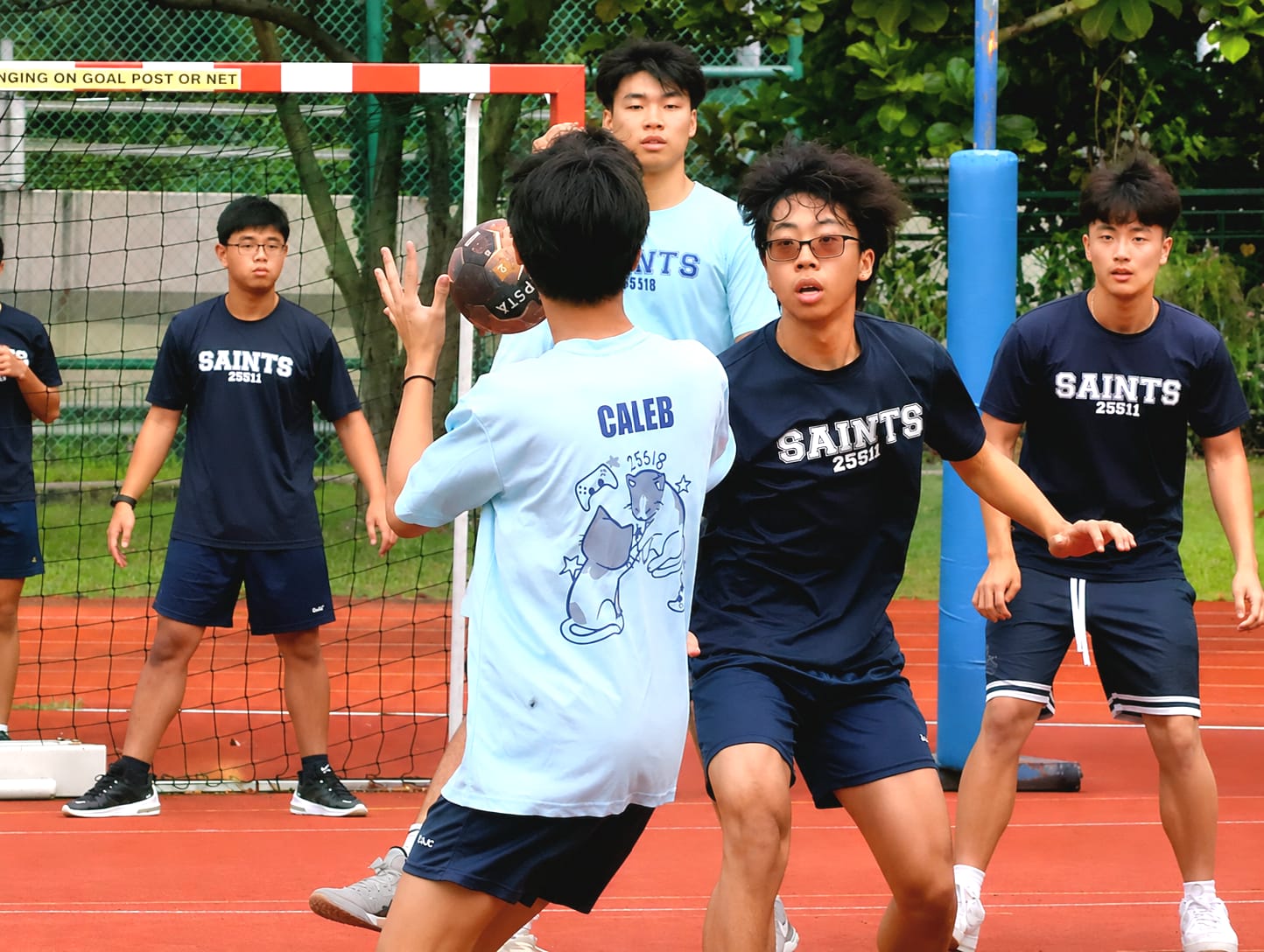 Two teams of students engaged in a friendly handball match, with a boy in dark blue t-shirt defending against another in light blue t-shirt.