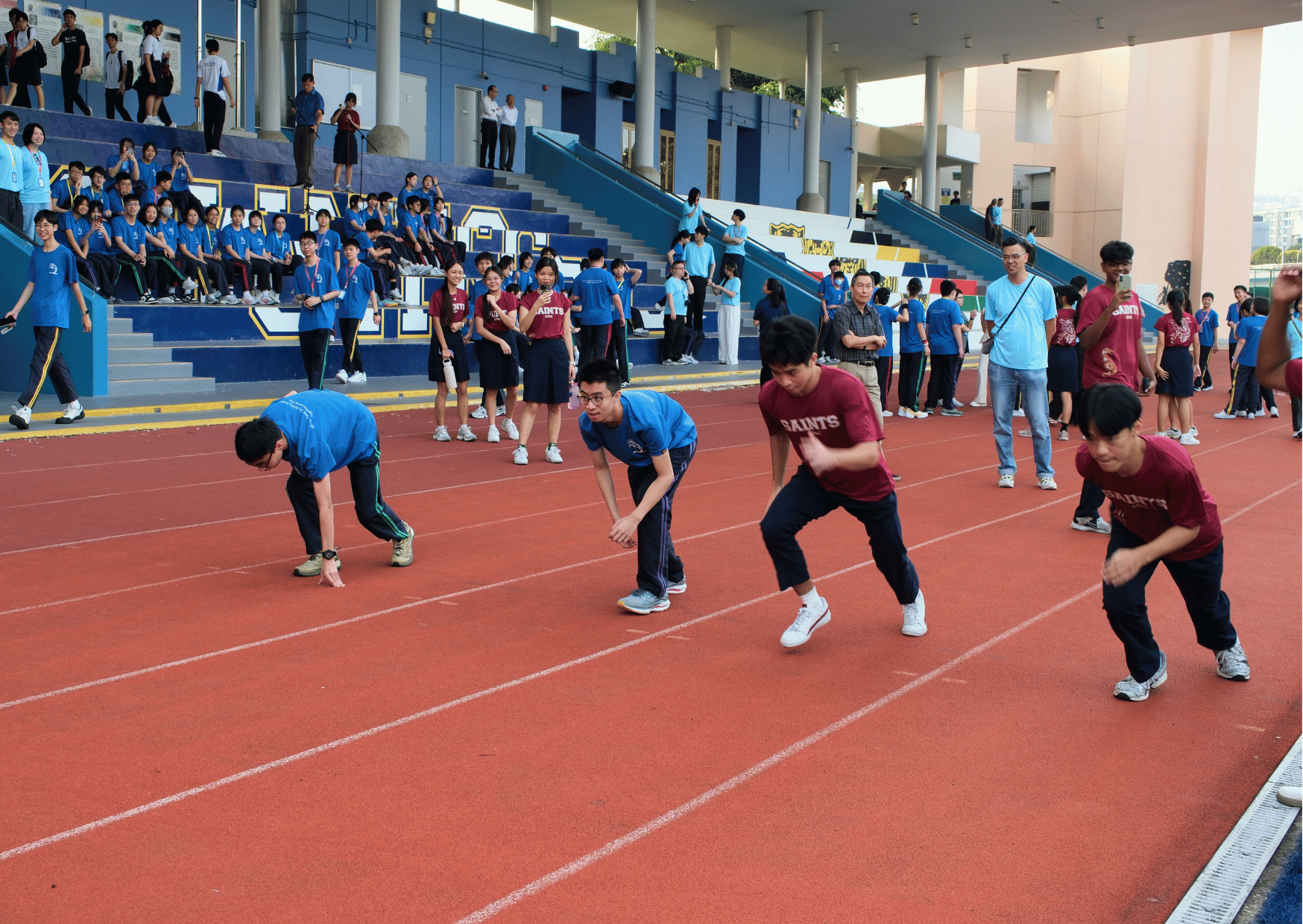 Students in blue and maroon "Saints" T-shirts crouch at the starting line of a running track, ready to race, while a crowd of fellow participants watches from the stands and sidelines.