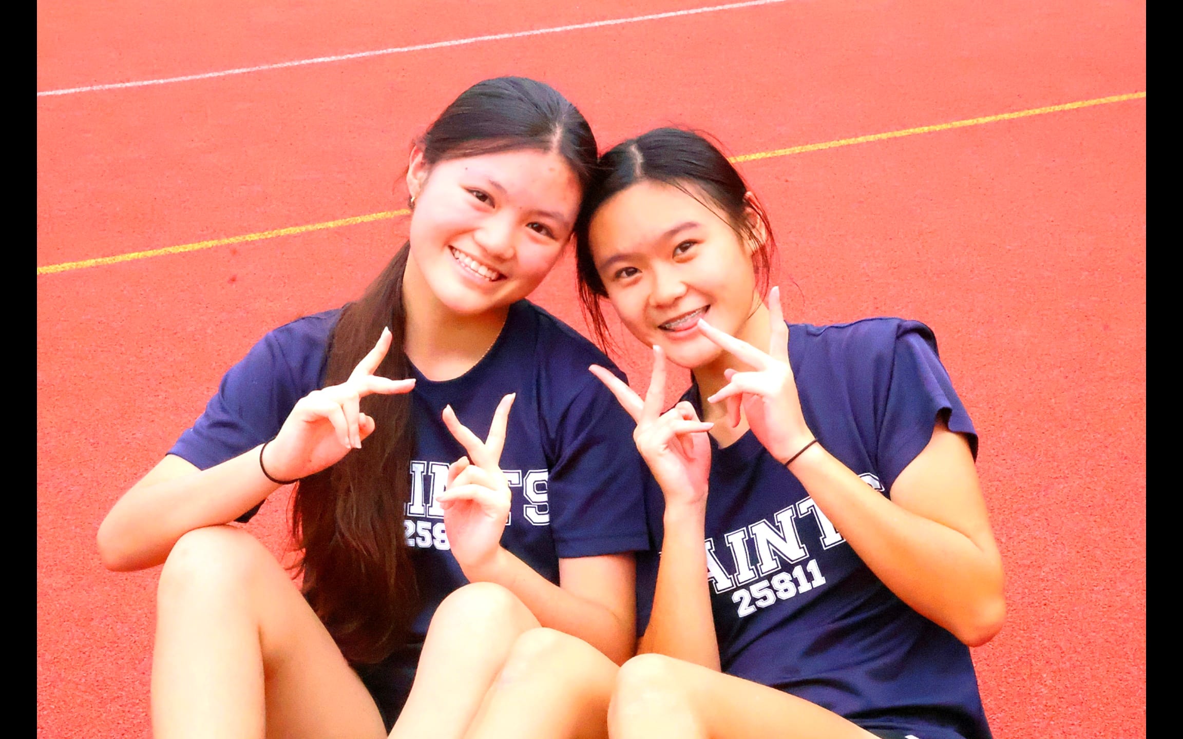 Two students seated on the track, smiling and posing for a shot for the camera. Both students holding up victory signs in both hands.