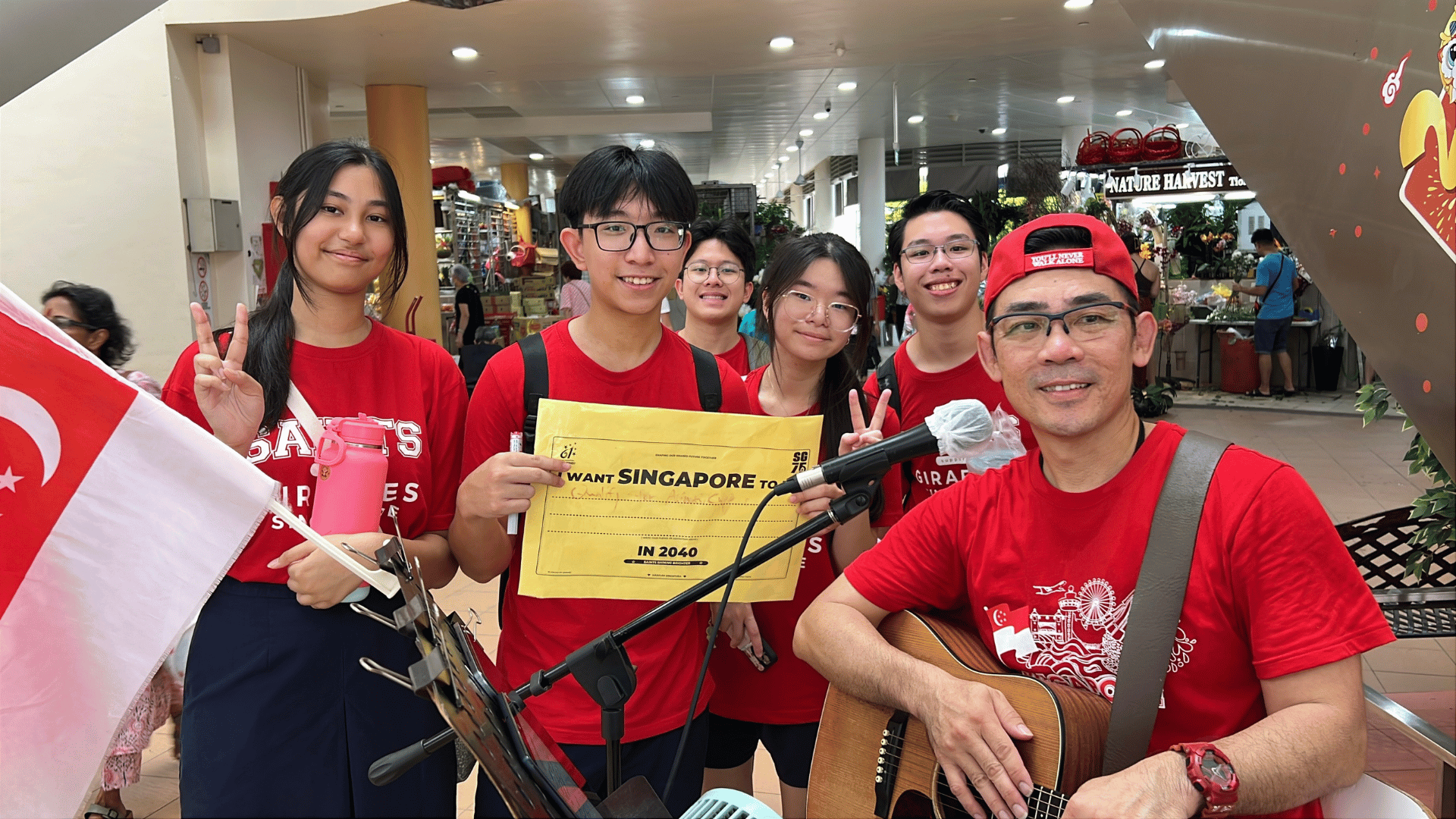 In an indoor public space, a man in a red baseball cap and red t-shirt sits in the foreground playing an acoustic guitar into a microphone. Beside him stand five students in matching red "SAINTS" t-shirts smiling for the camera, with one student holding up a yellow paper sign that reads "I WANT SINGAPORE TO BE... IN 2040." A small Singapore flag is partially visible in the lower-left corner.