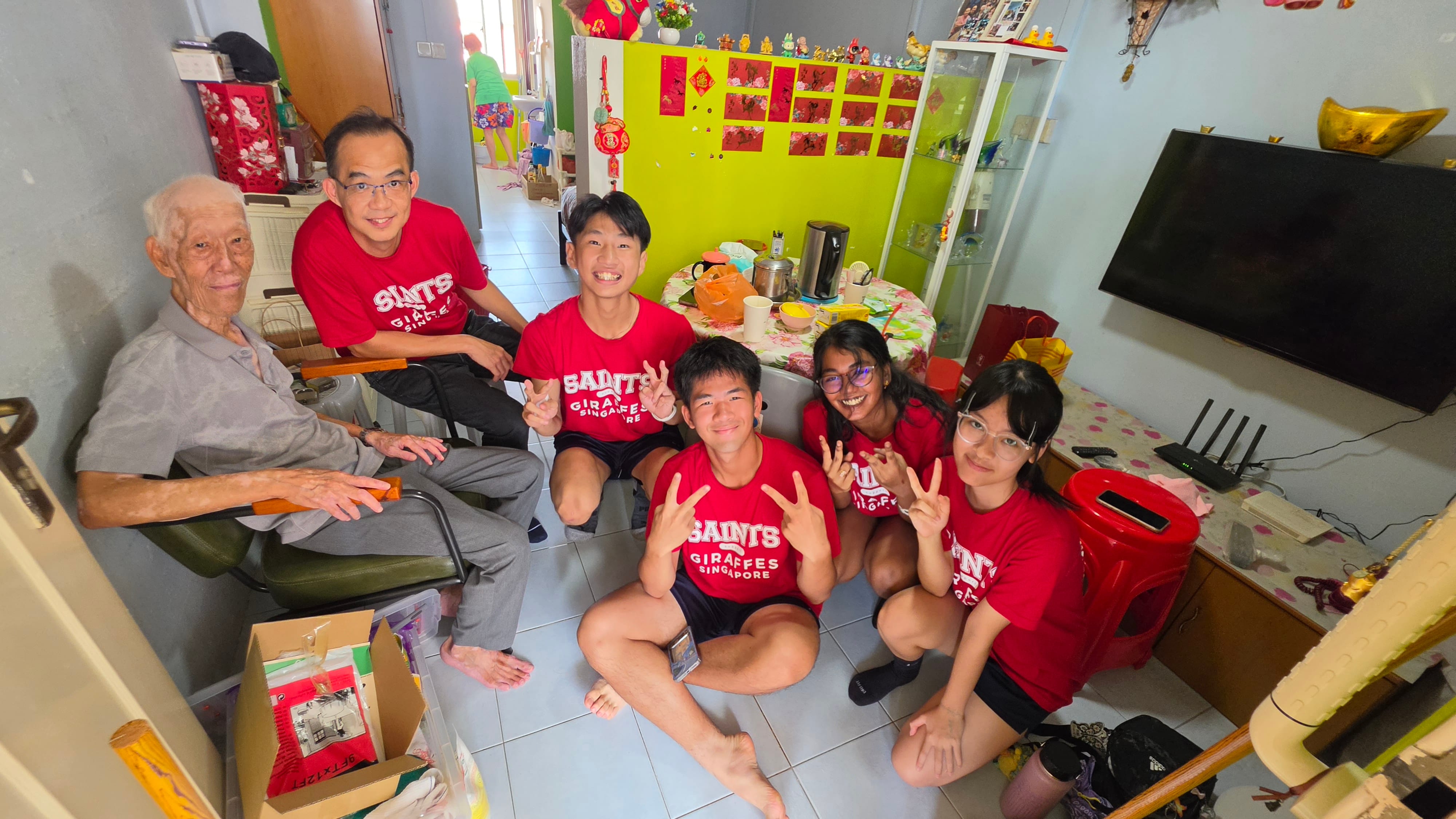 An indoor photo inside a home showing an elderly man seated in a green armchair next to five volunteers dressed in red "SAINTS GIRAFFES SINGAPORE" t-shirts. Four young student volunteers sit on the tiled floor smiling and giving peace signs, while an adult leader kneels beside the resident. In the background, a bright lime green partition wall decorated with red Chinese calligraphy couplets sits beside a mounted flat-screen TV and glass display cabinet.