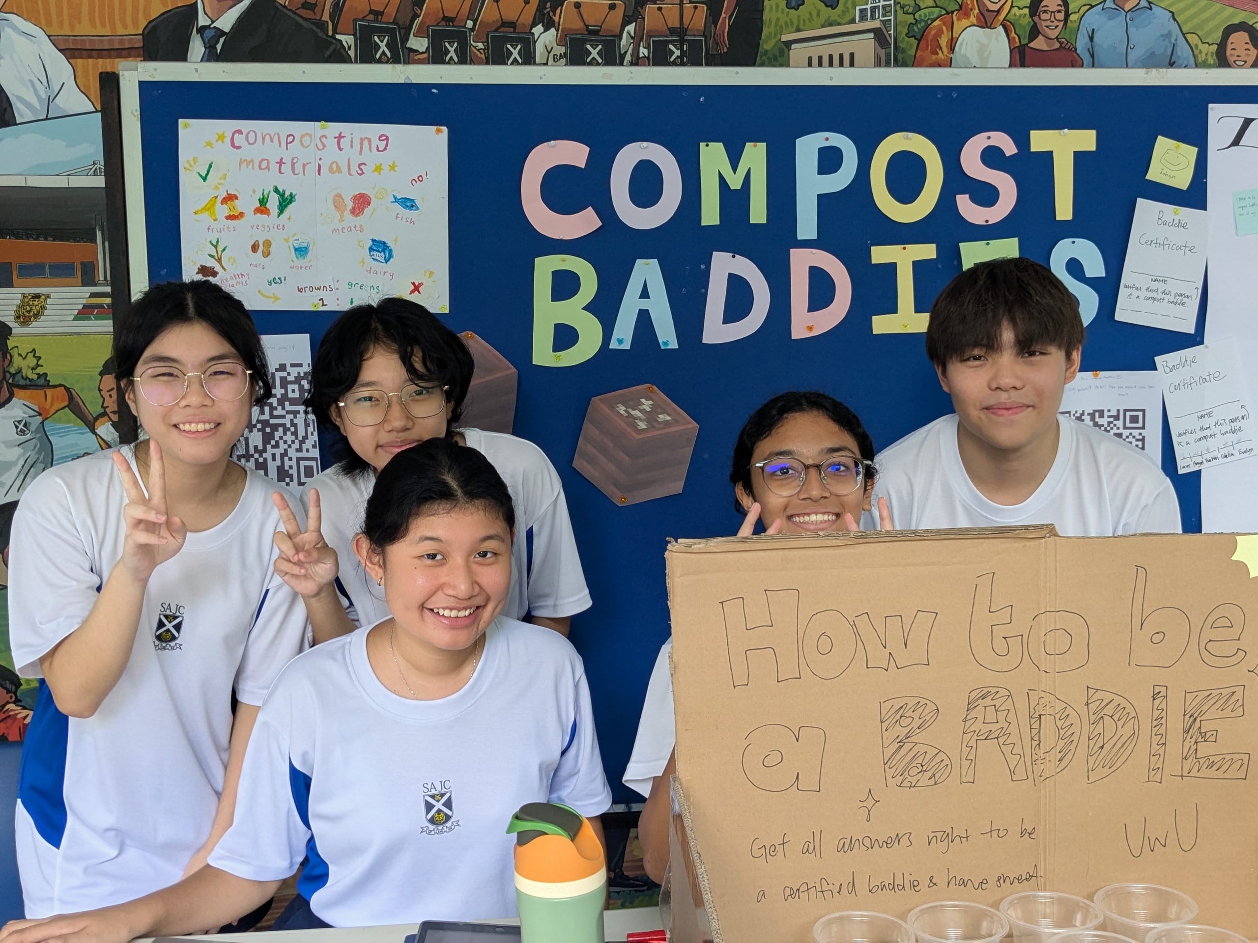 Five smiling students in white uniforms pose at a Compost Baddies booth with a cardboard sign reading How to be a Baddie.