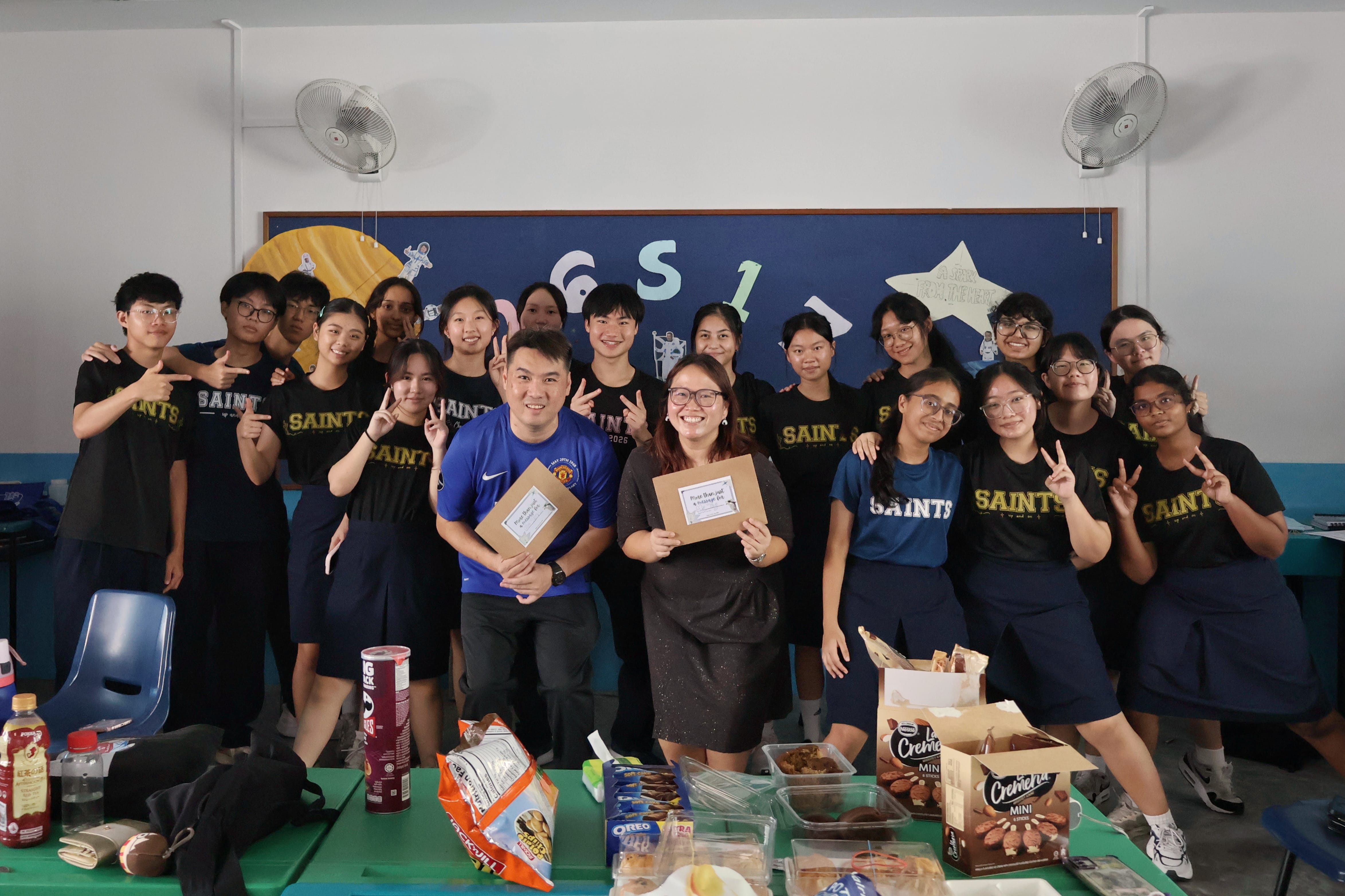 A cheerful group photo of students wearing dark "SAINTS" t-shirts gathered around two adults in the center who are holding brown gift envelopes. In the foreground, a table is filled with party snacks, chips, and boxed ice cream treats. A dark blue bulletin board hangs on the wall behind the group between two wall-mounted electric fans.