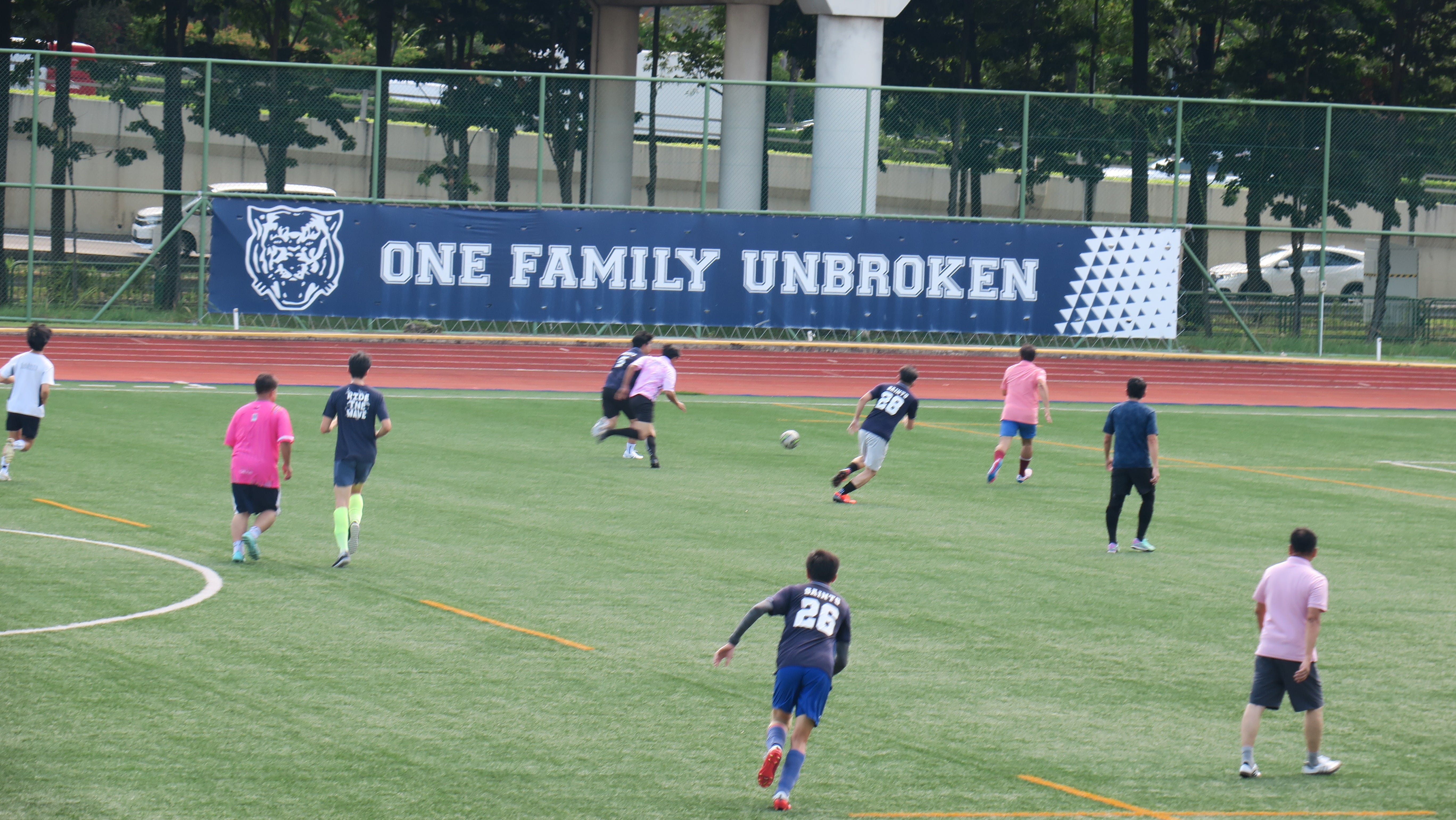 Players in pink and navy jerseys compete in a football match on a green pitch, with a large banner reading "One Family Unbroken" displayed along the perimeter fence.