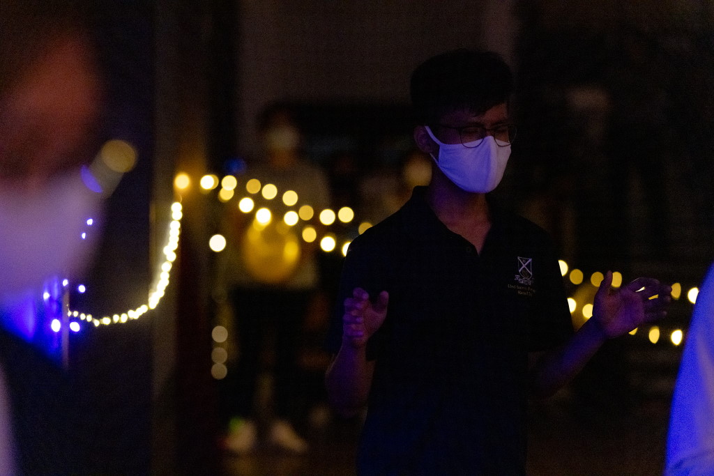 A student wearing a face mask and glasses stands with hands clasped in a dimly lit space decorated with warm string lights in the background.