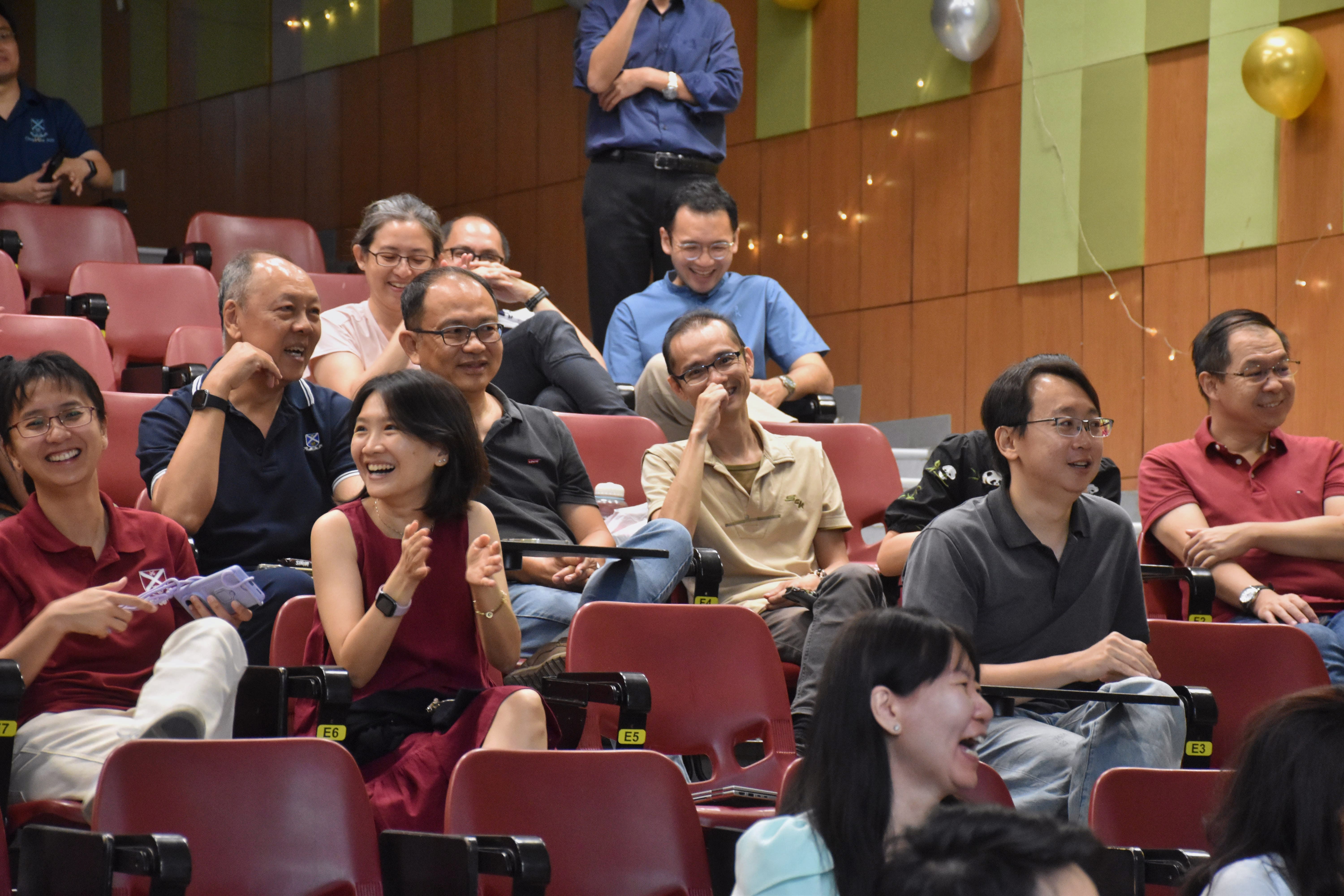 A cheerful audience of men and women seated in tiered red auditorium chairs. In the foreground, a woman in a maroon sleeveless dress claps enthusiastically alongside smiling colleagues. The people behind her are laughing and watching a presentation in a wood-and-green paneled lecture hall decorated with festive balloons.