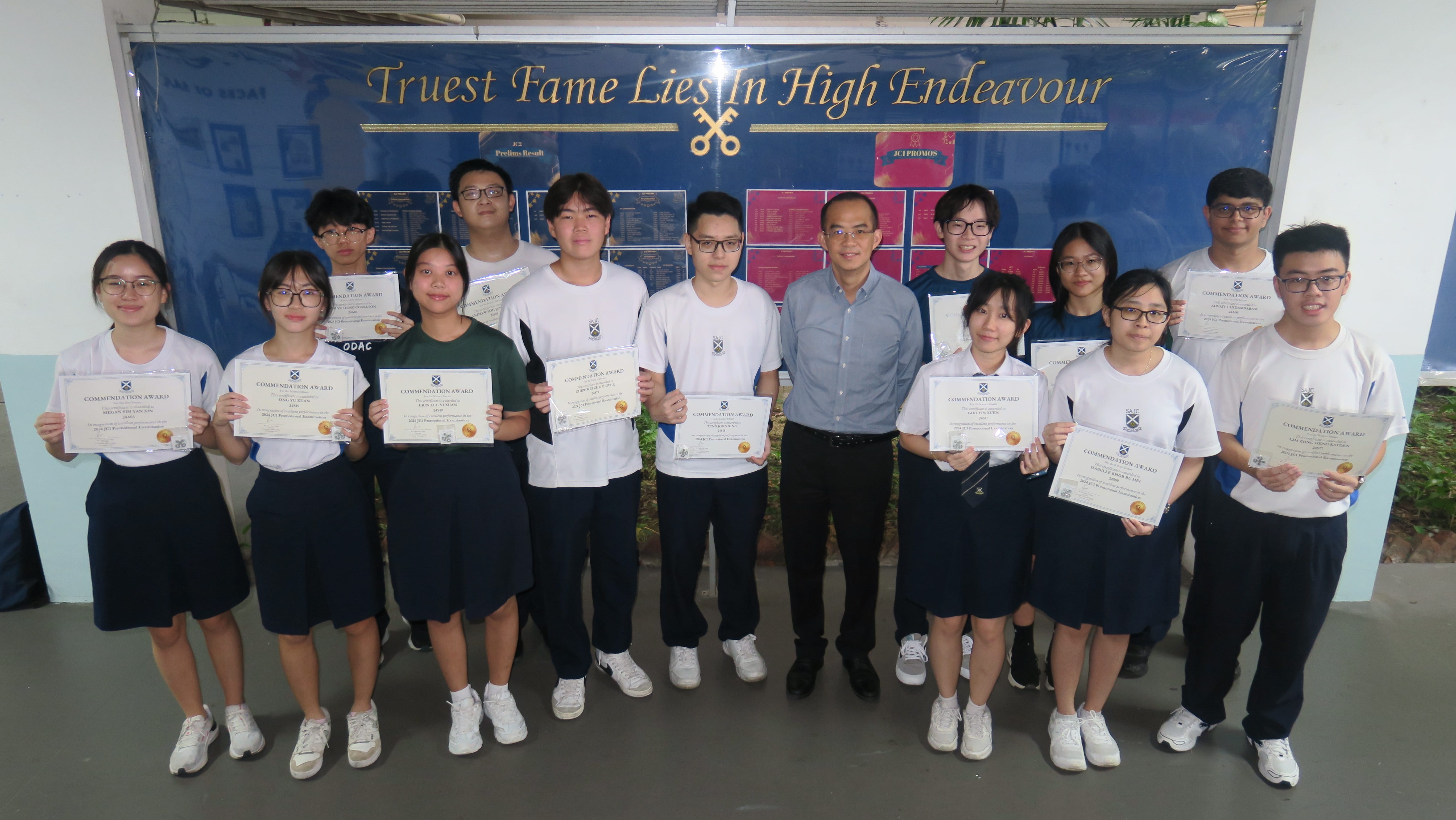 Students and a teacher pose holding Commendation Award certificates in front of a blue school notice board.