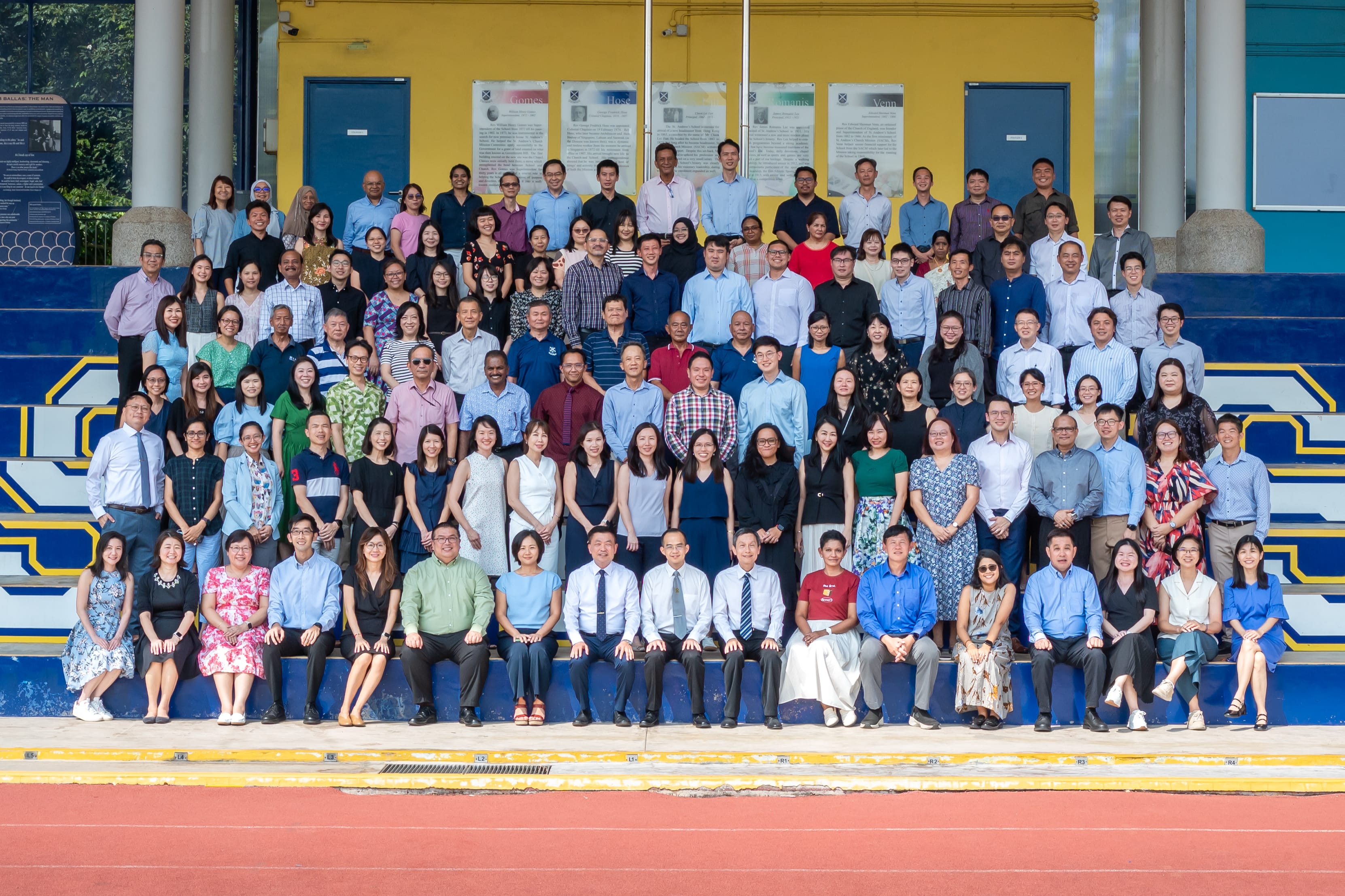 Large group photo of many people on blue stadium steps in front of a yellow wall with posters and doors.