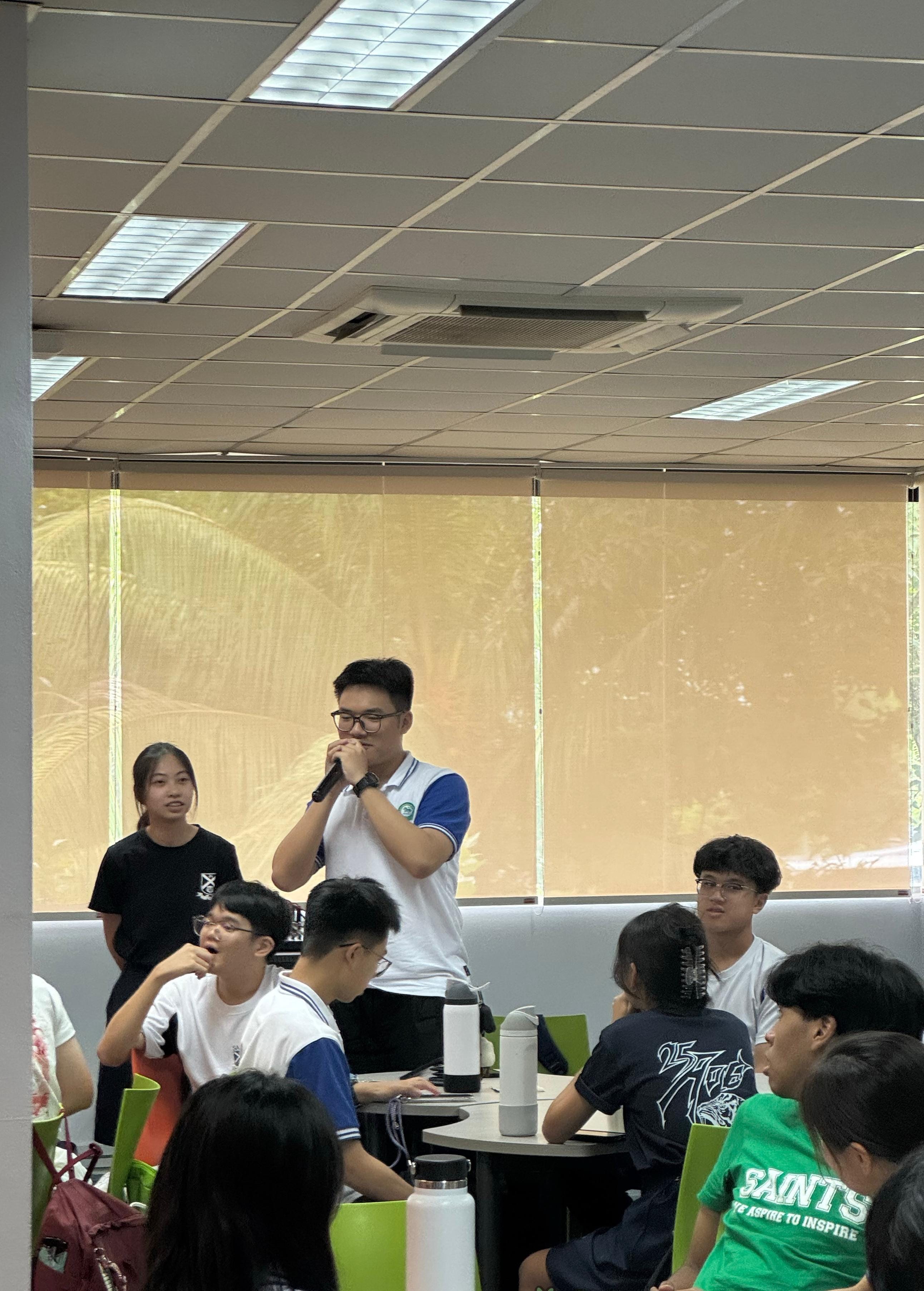 A visiting student from Hangzhou speaks into a microphone while standing in a classroom, addressing a mixed group of local and visiting students during an exchange interaction session at St Andrew's Junior College.