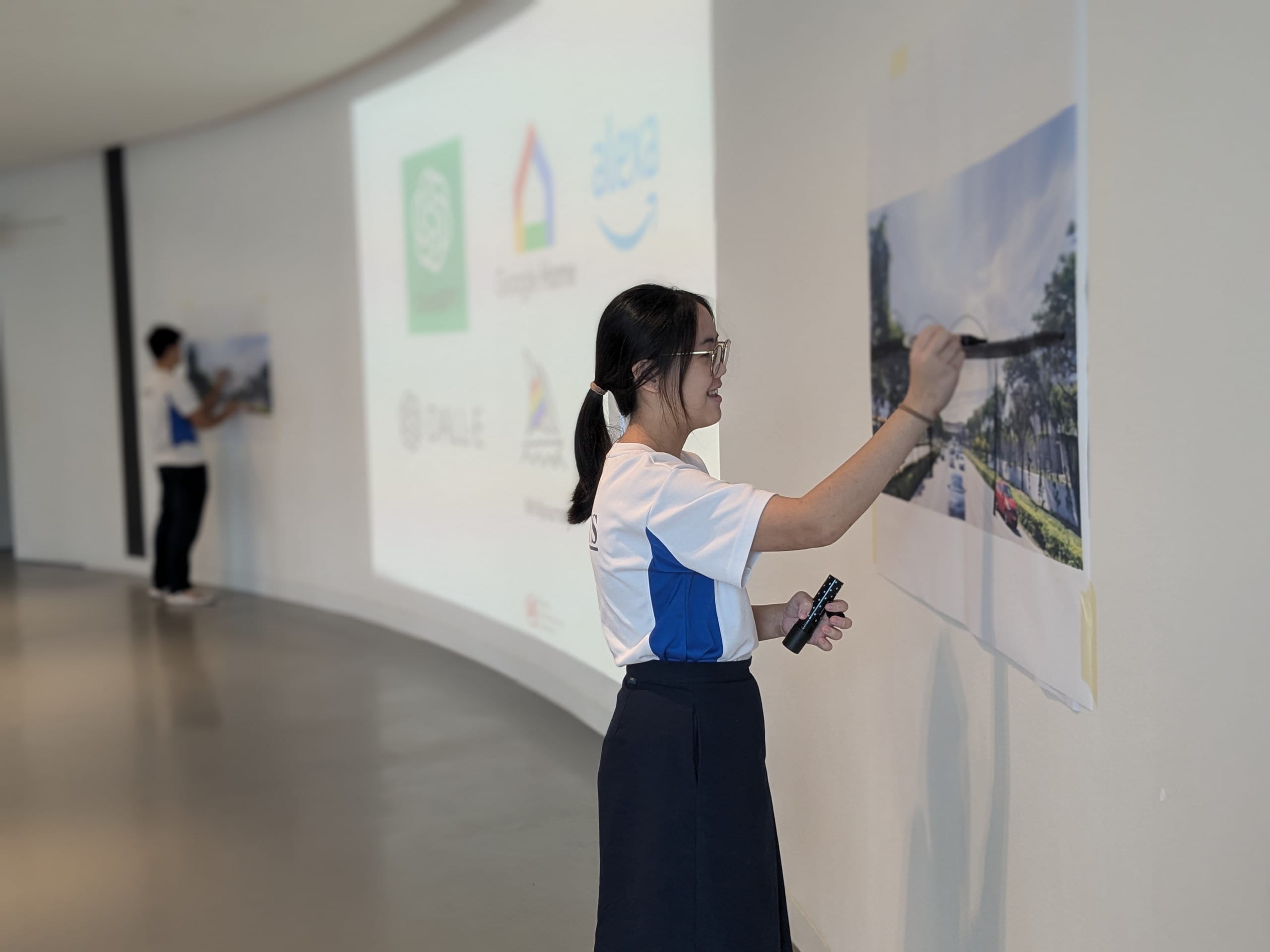 Young woman with glasses pinning a street scene photo to a white wall, with a projected presentation visible behind her.