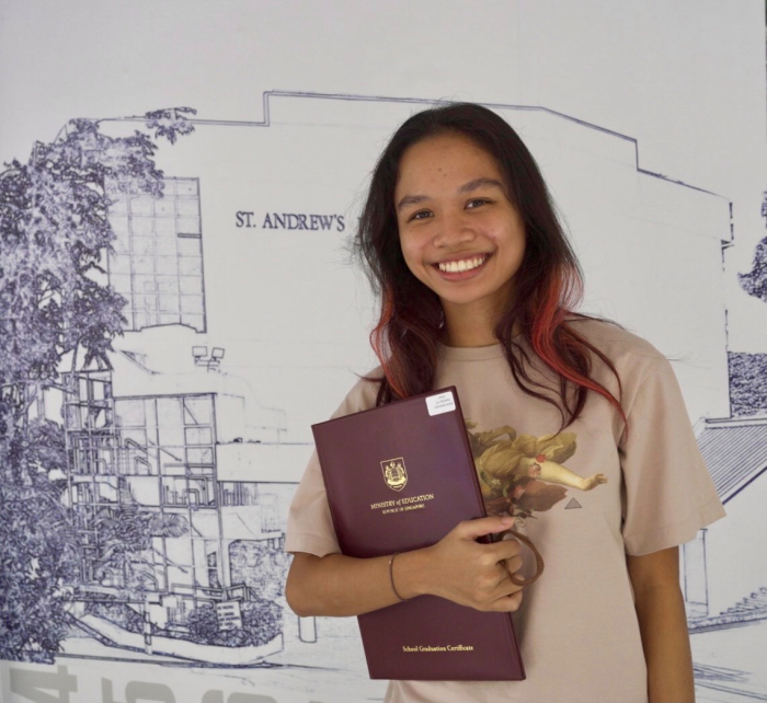 Young woman with long red-tipped hair smiling, holding a School Graduation Certificate folder in front of a St. Andrew's mural.