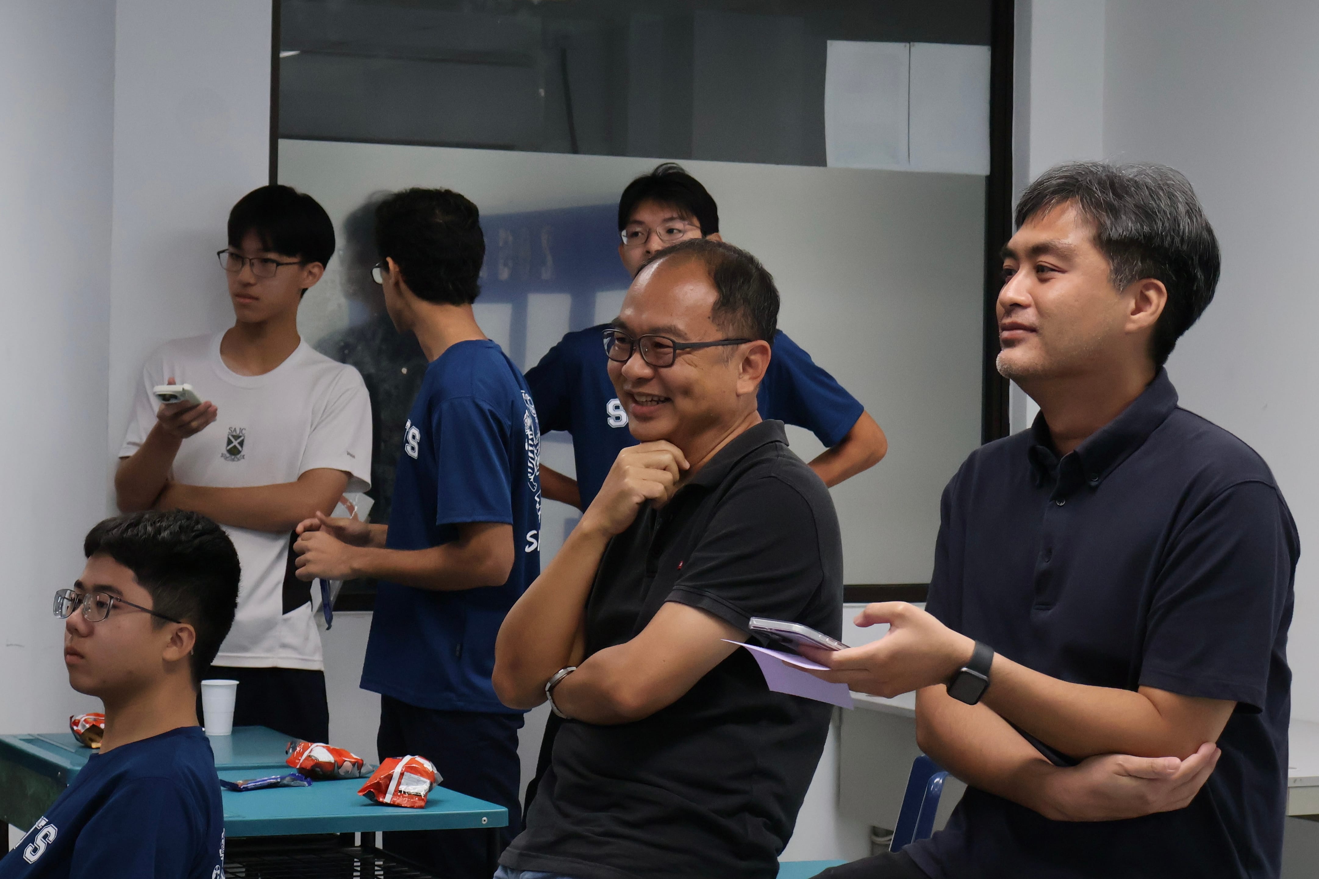 Two men in dark polo shirts sit side-by-side, watching an off-camera event with amused and focused expressions. The man on the left wears glasses and rests a hand on his chin with a smile. In the background, several students in blue and white t-shirts stand near a glass partition, with snacks scattered on nearby tables.