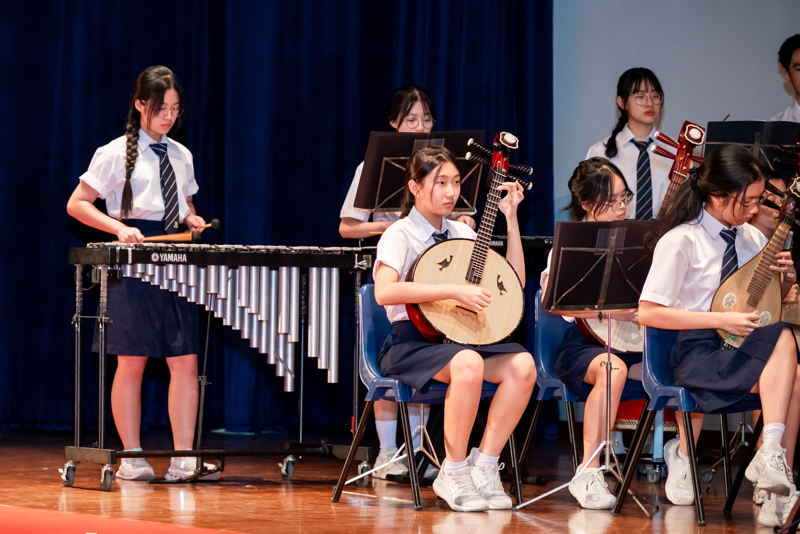 St Andrew's Junior College students in school uniform perform on stage with traditional Chinese instruments, including a ruan (Chinese lute) and pipa, alongside a Yamaha marimba, during a musical performance.