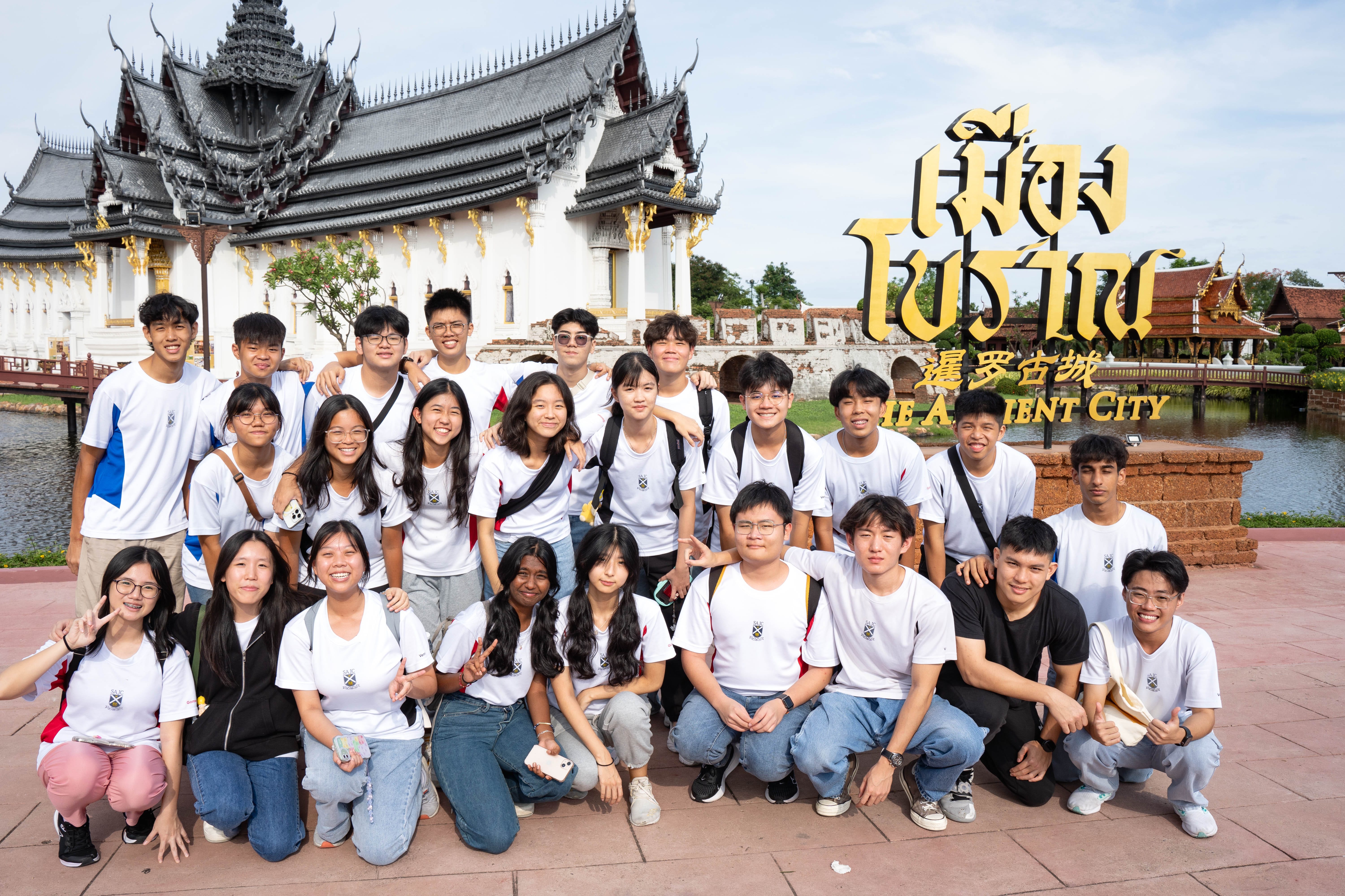Students from St Andrew's Junior College in white college T-shirts pose for a group photo at the Ancient City (Mueang Boran) attraction in Bangkok, with a traditional Thai temple and the landmark's gold signage visible in the background.