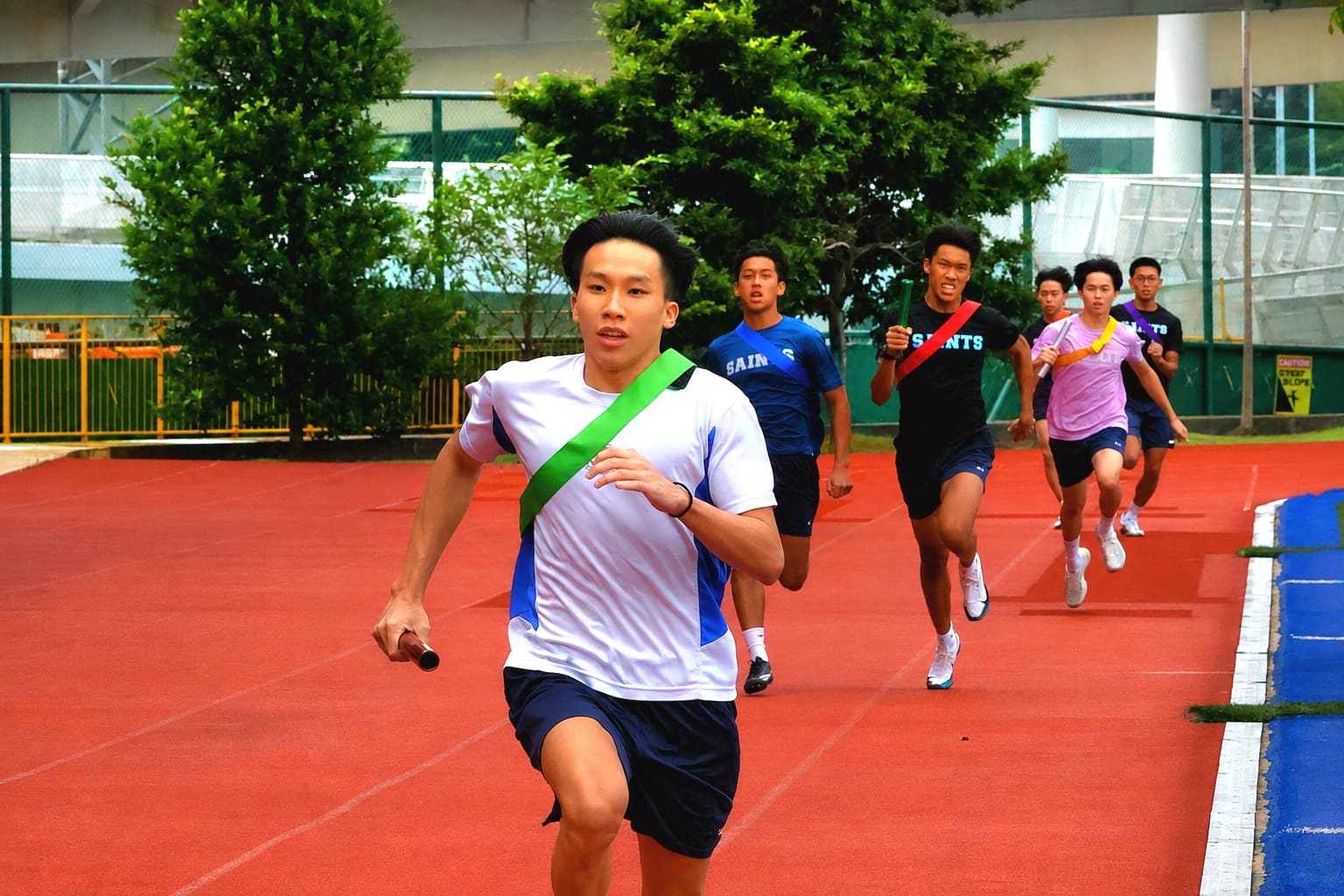 Male student athletes in colored sashes sprinting during a relay race on a red track, led by a runner in a white shirt holding a baton.