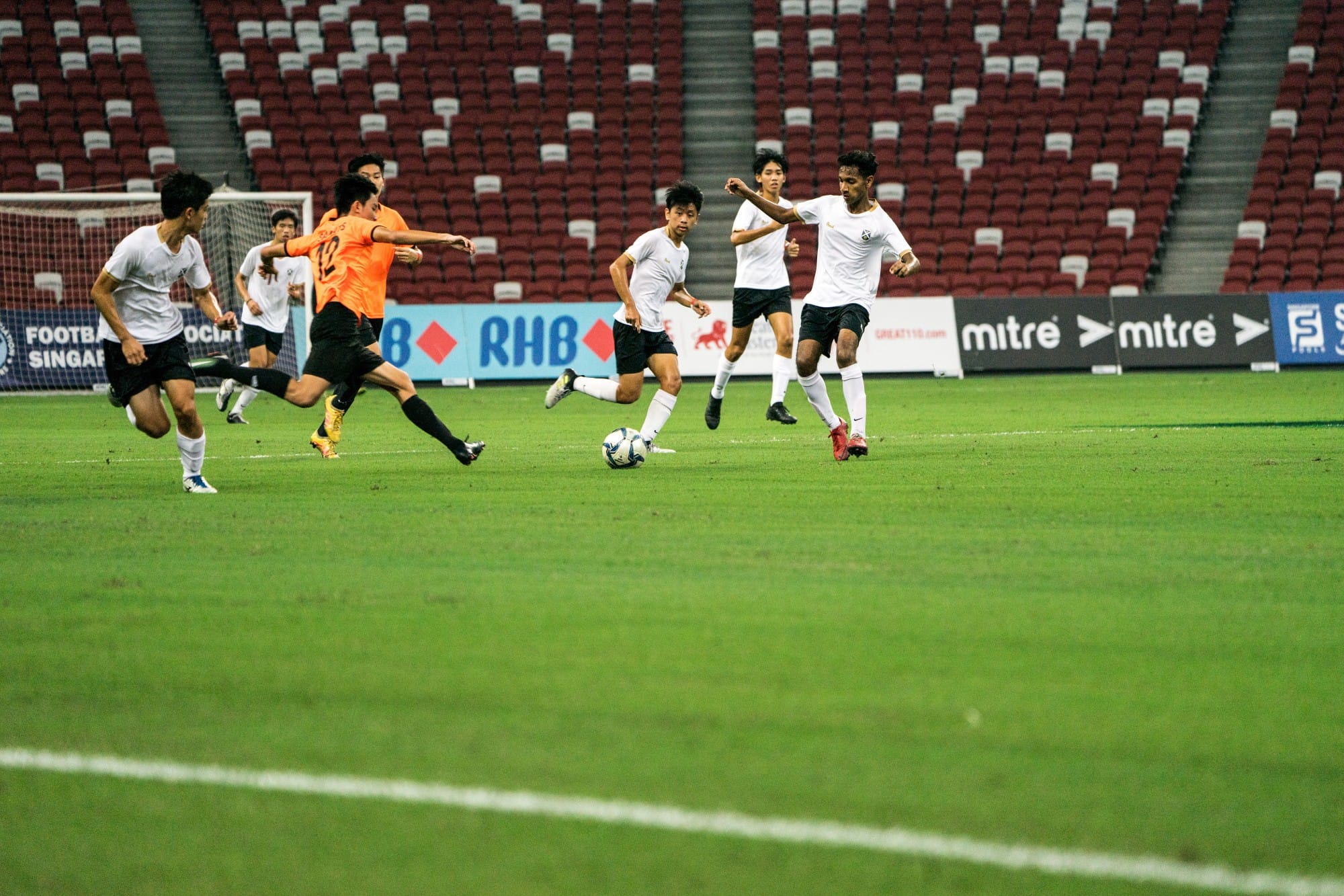 SAJC Boys' Football team in action during a match at a stadium, with a player in white dribbling past an opponent in orange