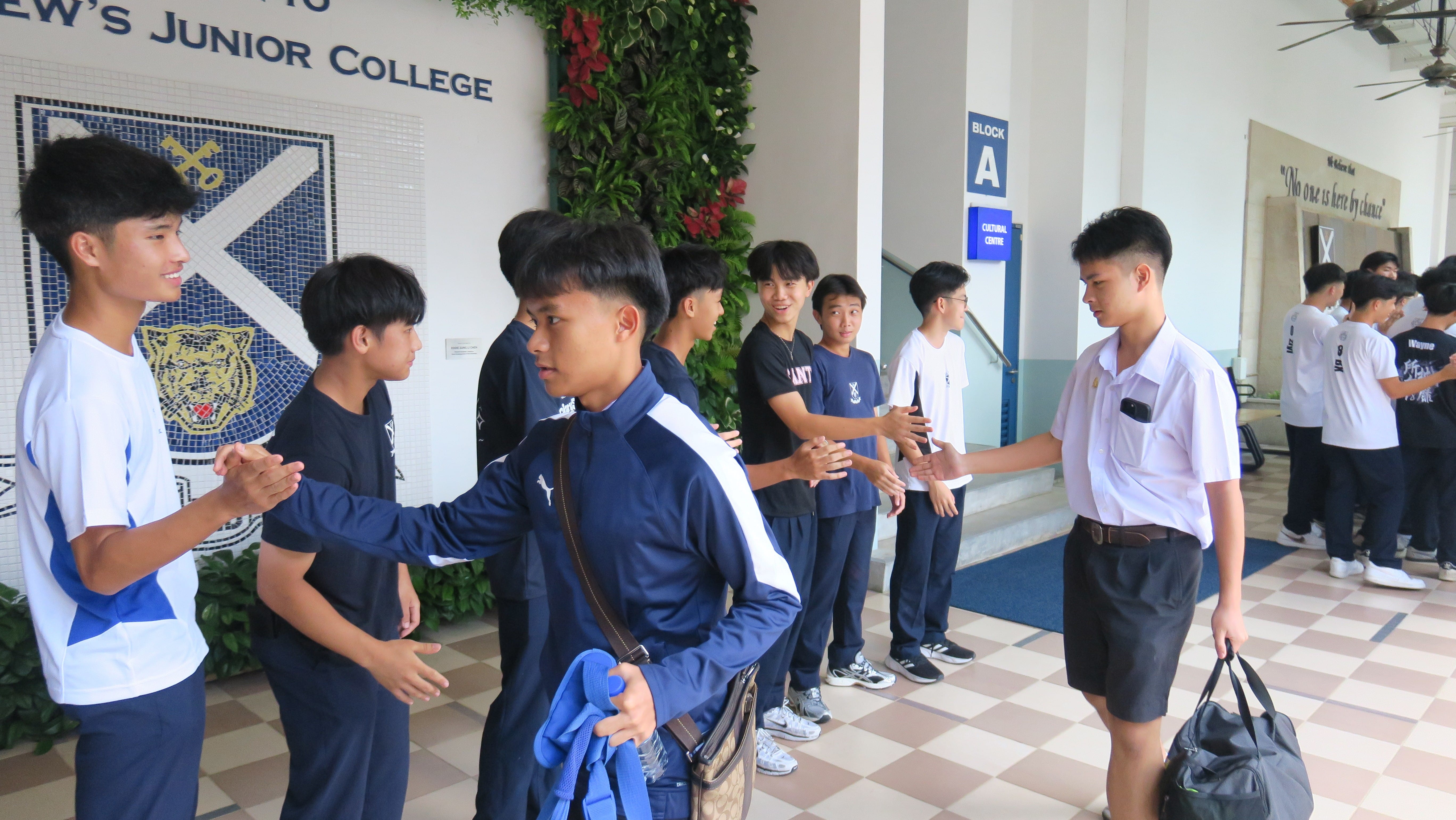 Visiting students are welcomed with handshakes by Saints students in the foyer of St Andrew's Junior College, with the school crest and motto visible on the wall behind them.